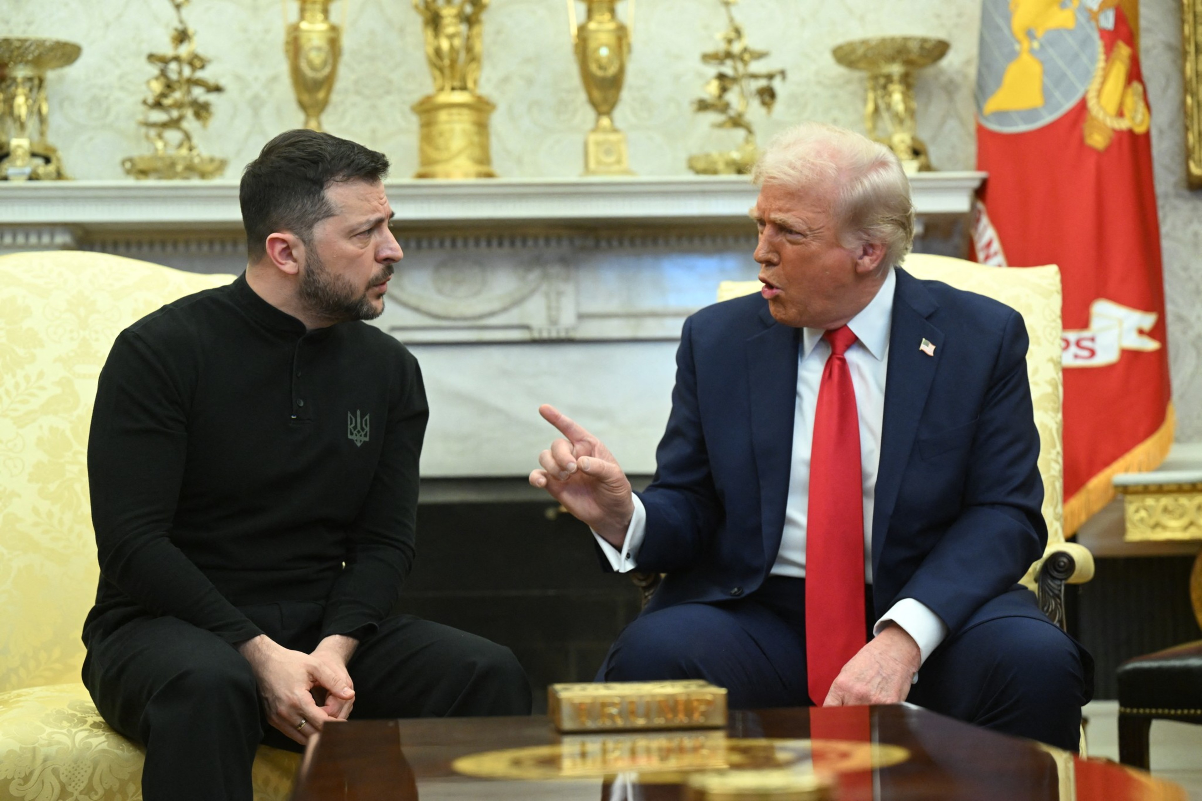 Two men are seated on yellow chairs in a formal room. The man on the left wears black, while the man on the right, in a blue suit and red tie, gestures emphatically.