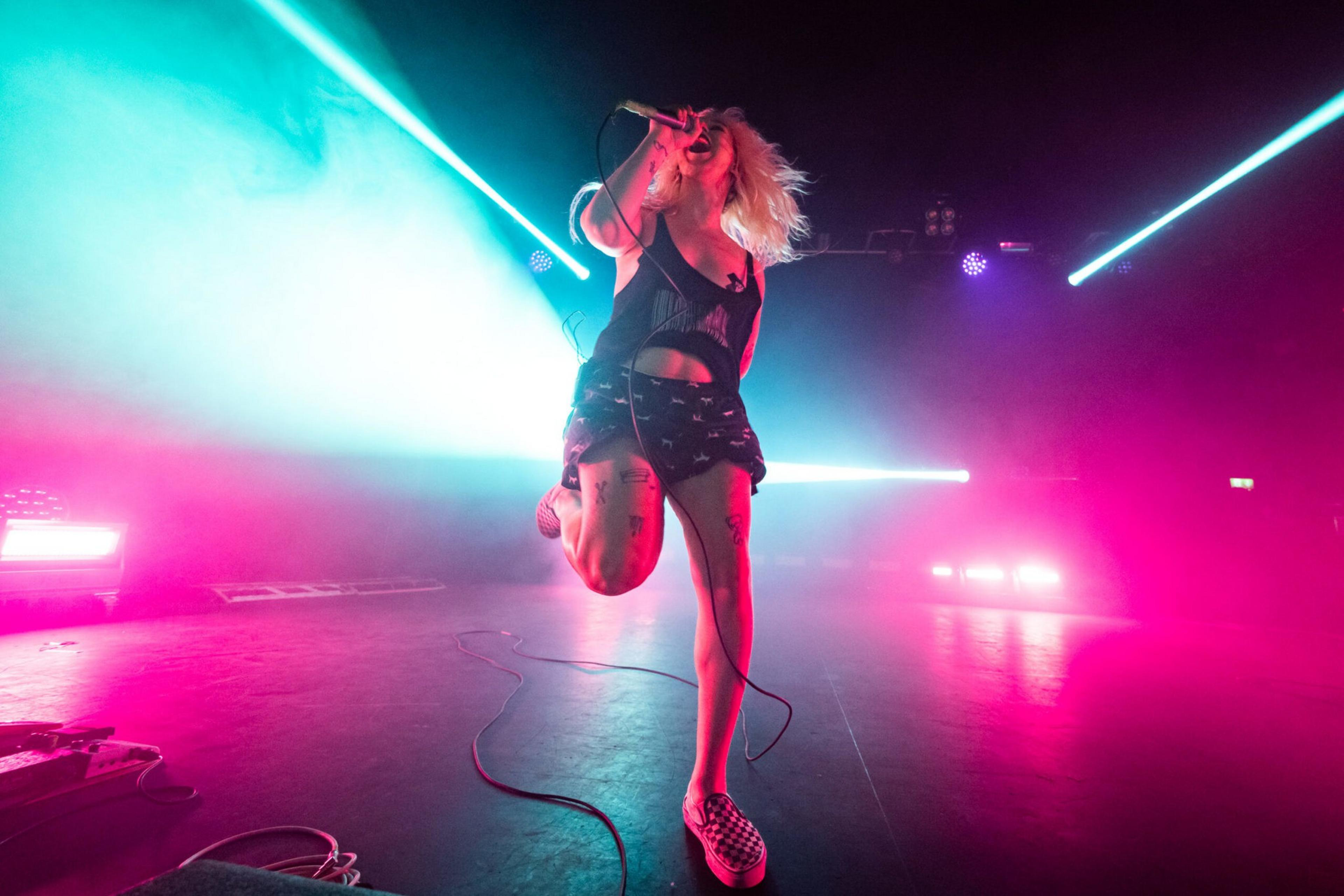 A performer with blonde hair passionately sings into a microphone on stage, surrounded by vibrant blue and pink lights and fog effects.