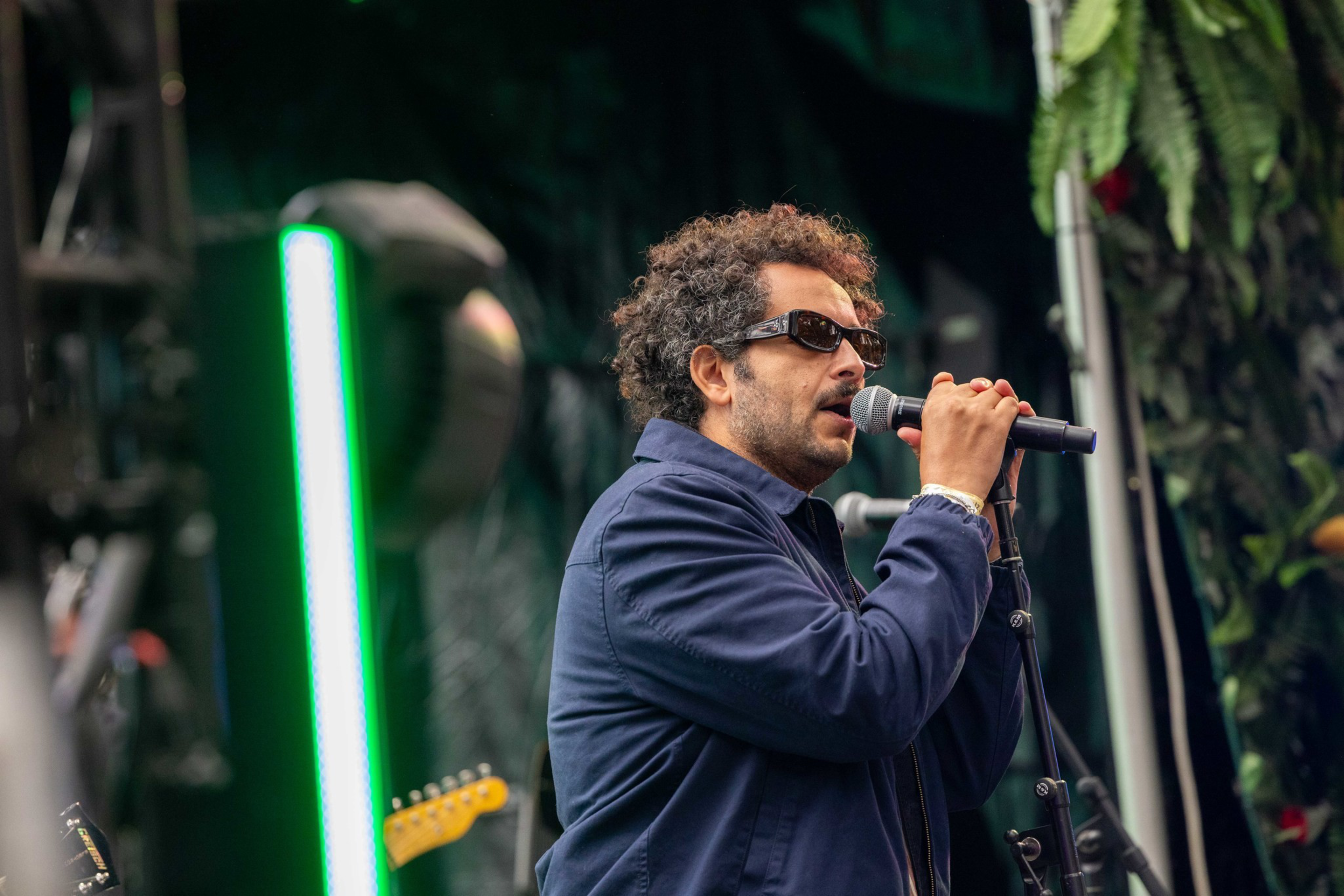 A man with curly hair and sunglasses is singing into a microphone on stage, wearing a dark jacket with a green light and guitar in the background.