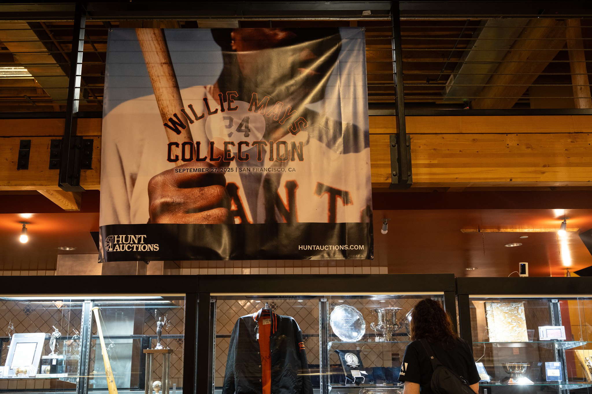 A person views a glass display case holding sports memorabilia including a jacket, trophies, plaques, and baseballs below a banner for Willie Mays Collection auction.