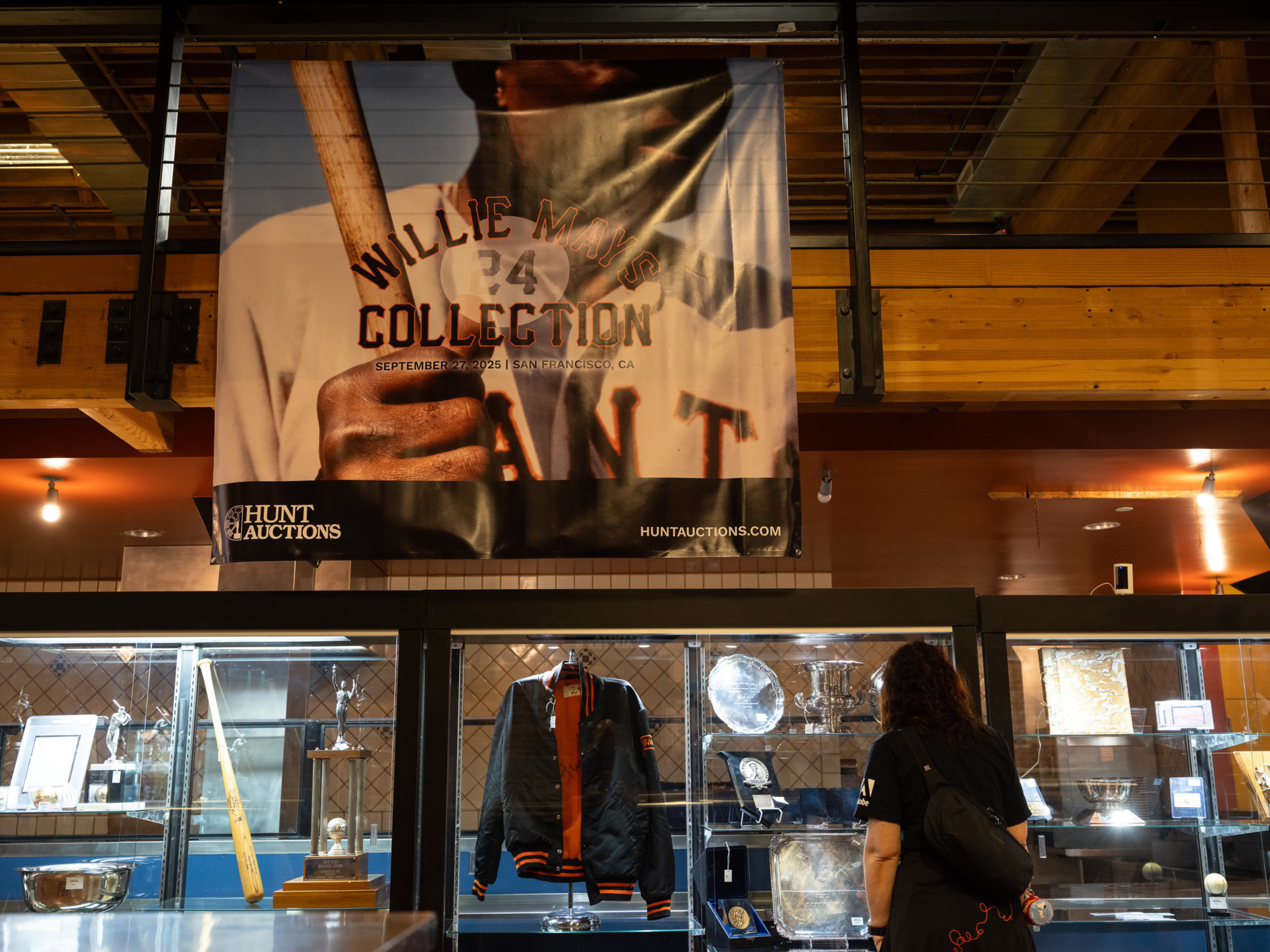 A person views a glass display case holding sports memorabilia including a jacket, trophies, plaques, and baseballs below a banner for Willie Mays Collection auction.
