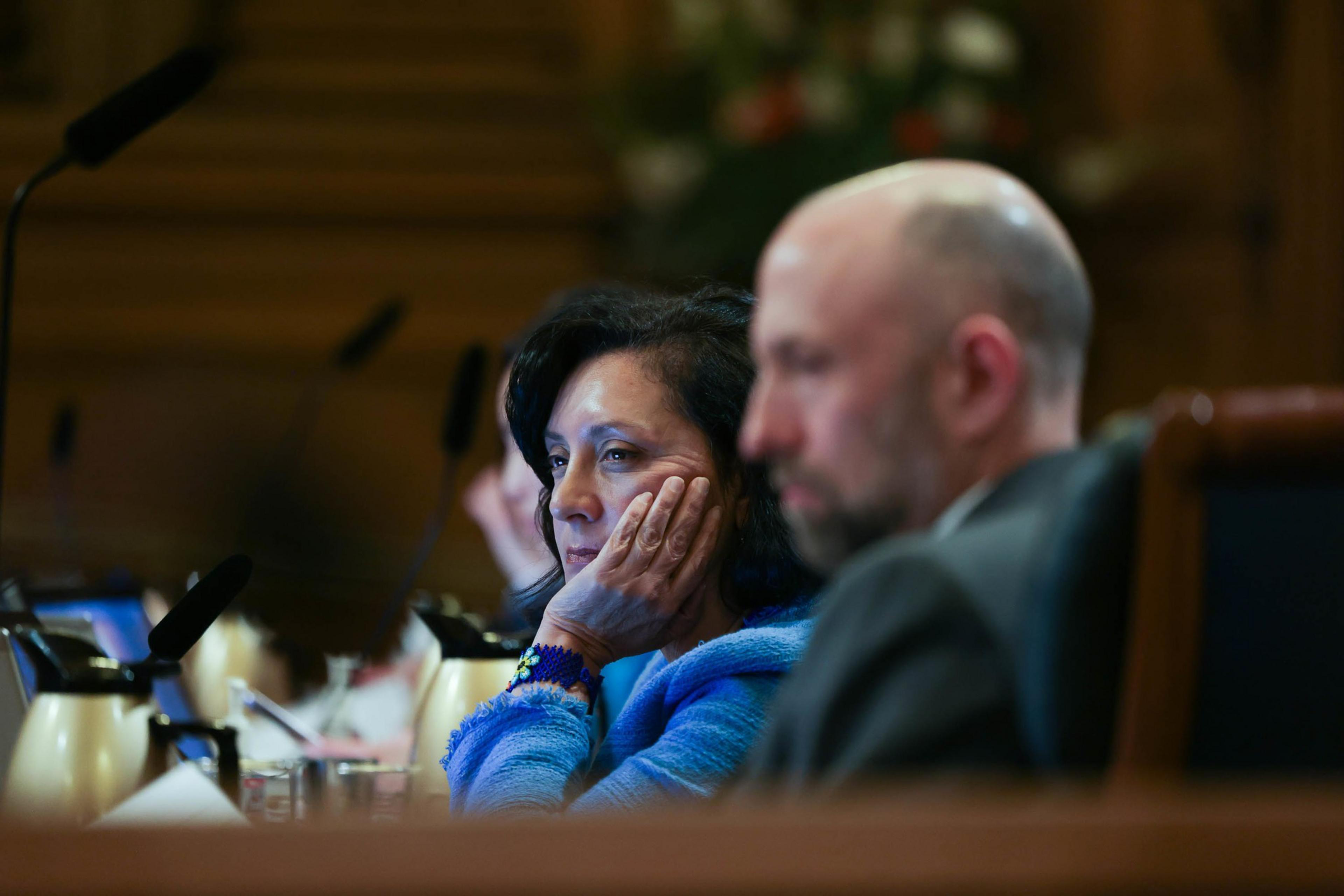 A woman in a blue sweater rests her head on her hand, looking thoughtful, while a man in a suit sits blurred in the foreground.