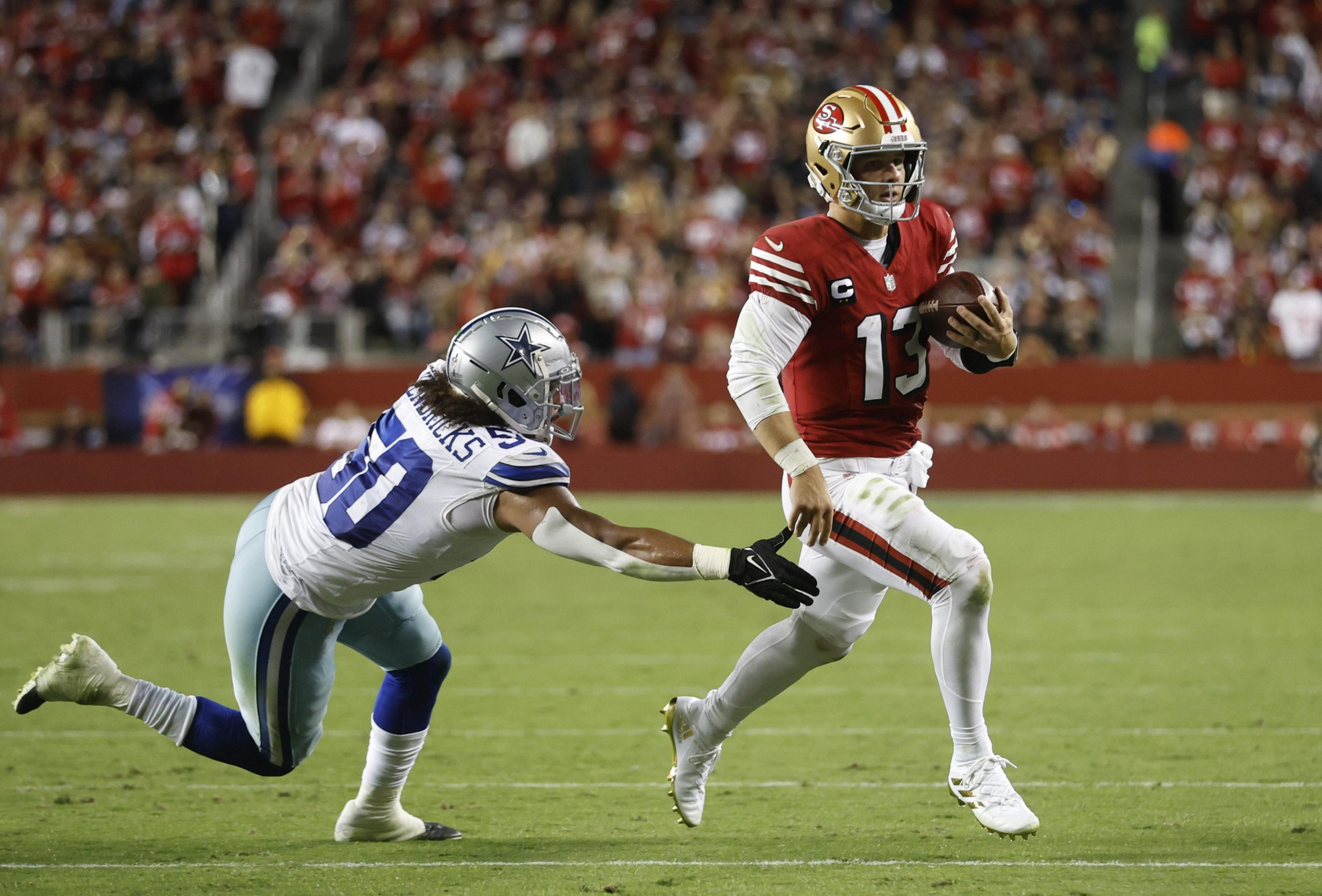 A football player in a red uniform is running with the ball as another player in a white and blue uniform reaches out, trying to tackle him on a grassy field.