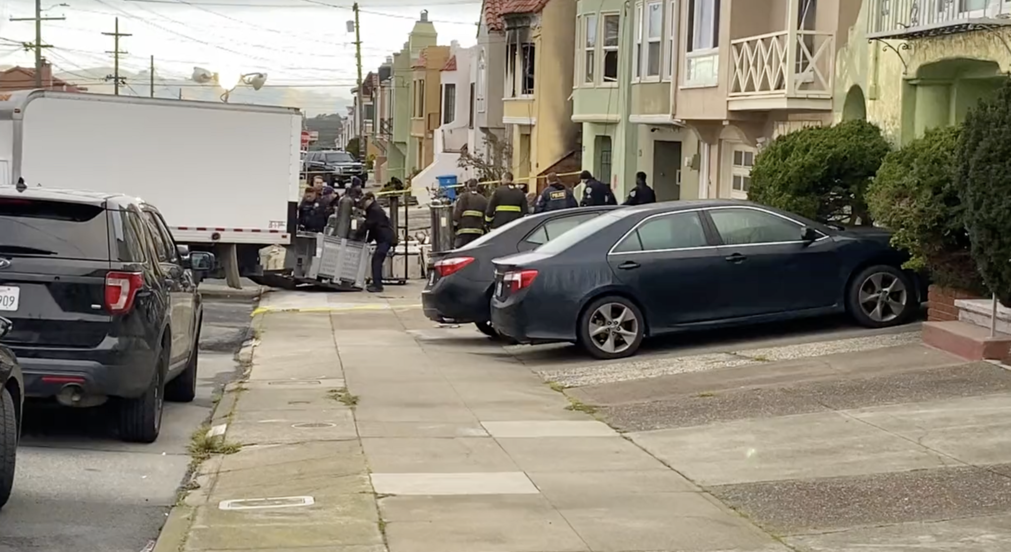 Emergency responders and police gather near a large white truck parked on a residential street with parked cars along the sidewalk.