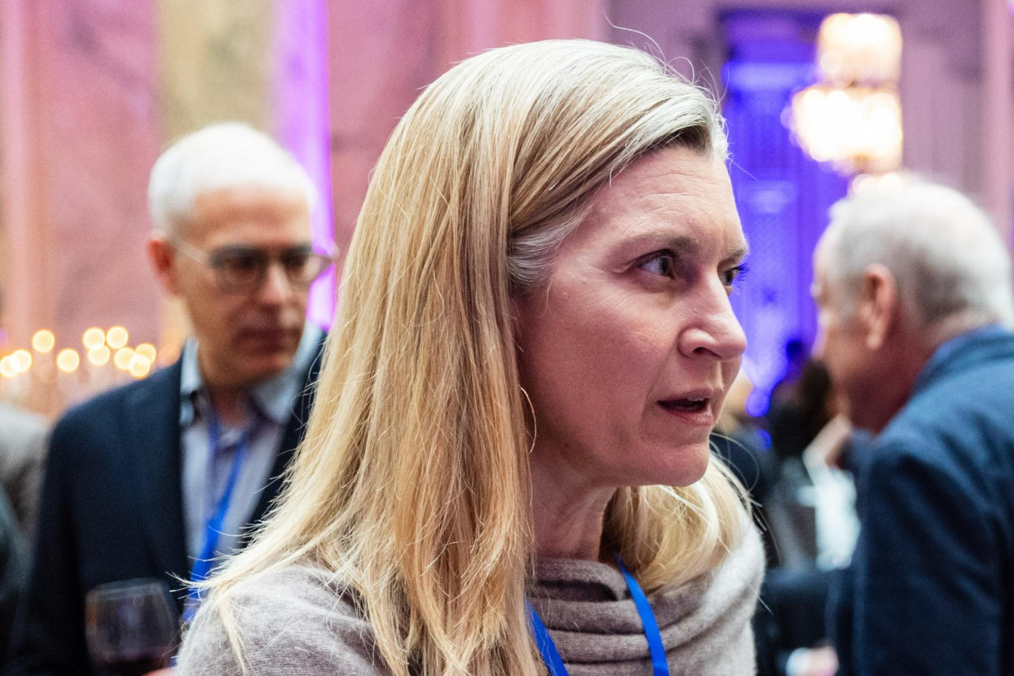 A woman is engaged in conversation at an event. She wears a light sweater and has a blue lanyard. People in the background are holding drinks, with dim, colorful lighting.