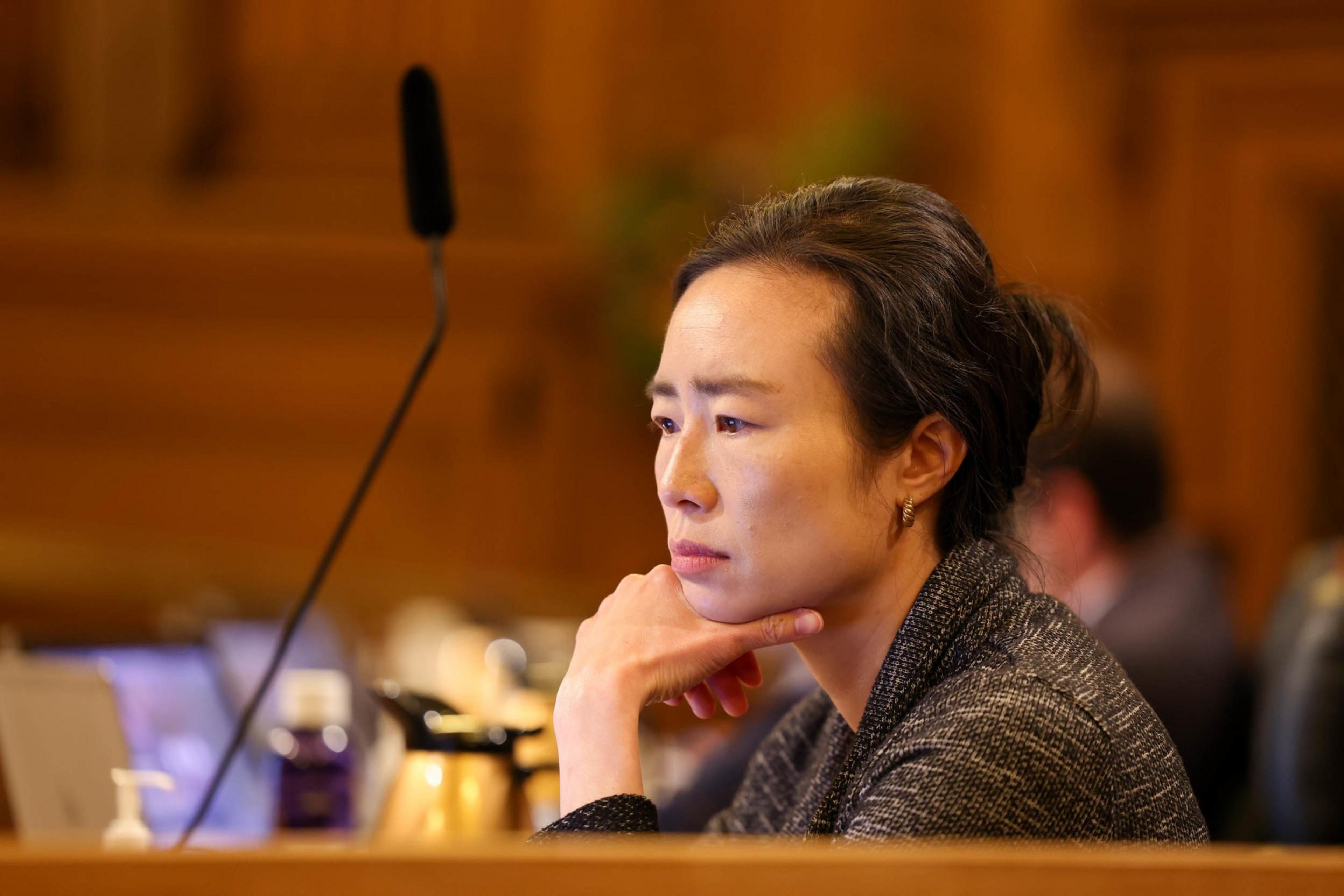 A person sits pensively with their chin resting on their hand. They appear engaged in thought. In the background are blurred figures and indoor lighting.