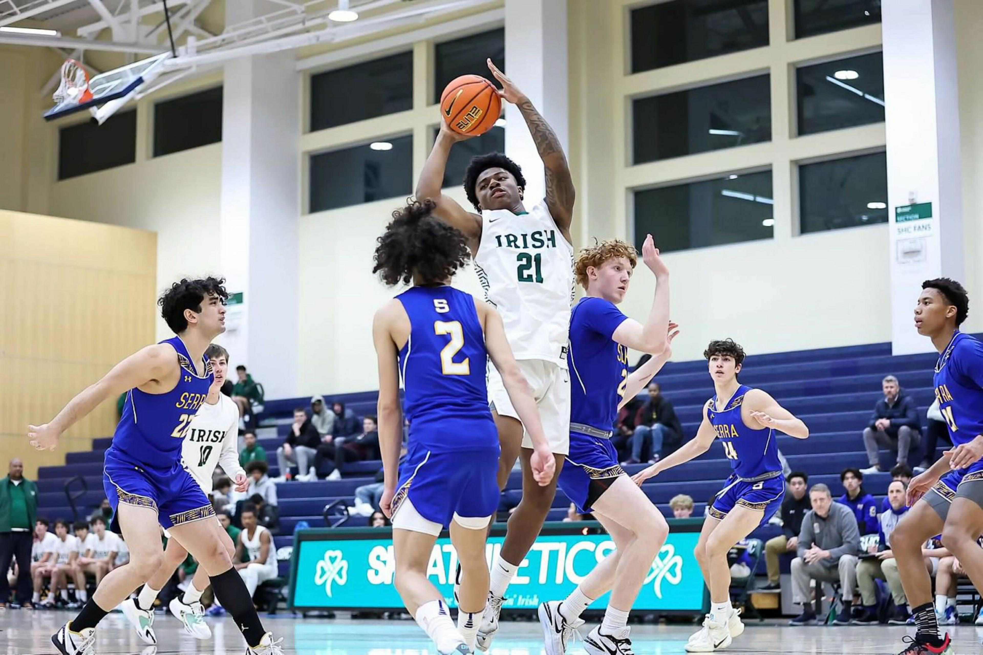 A basketball player in a white "Irish" jersey jumps to shoot while five players in blue jerseys try to block him inside a gym.