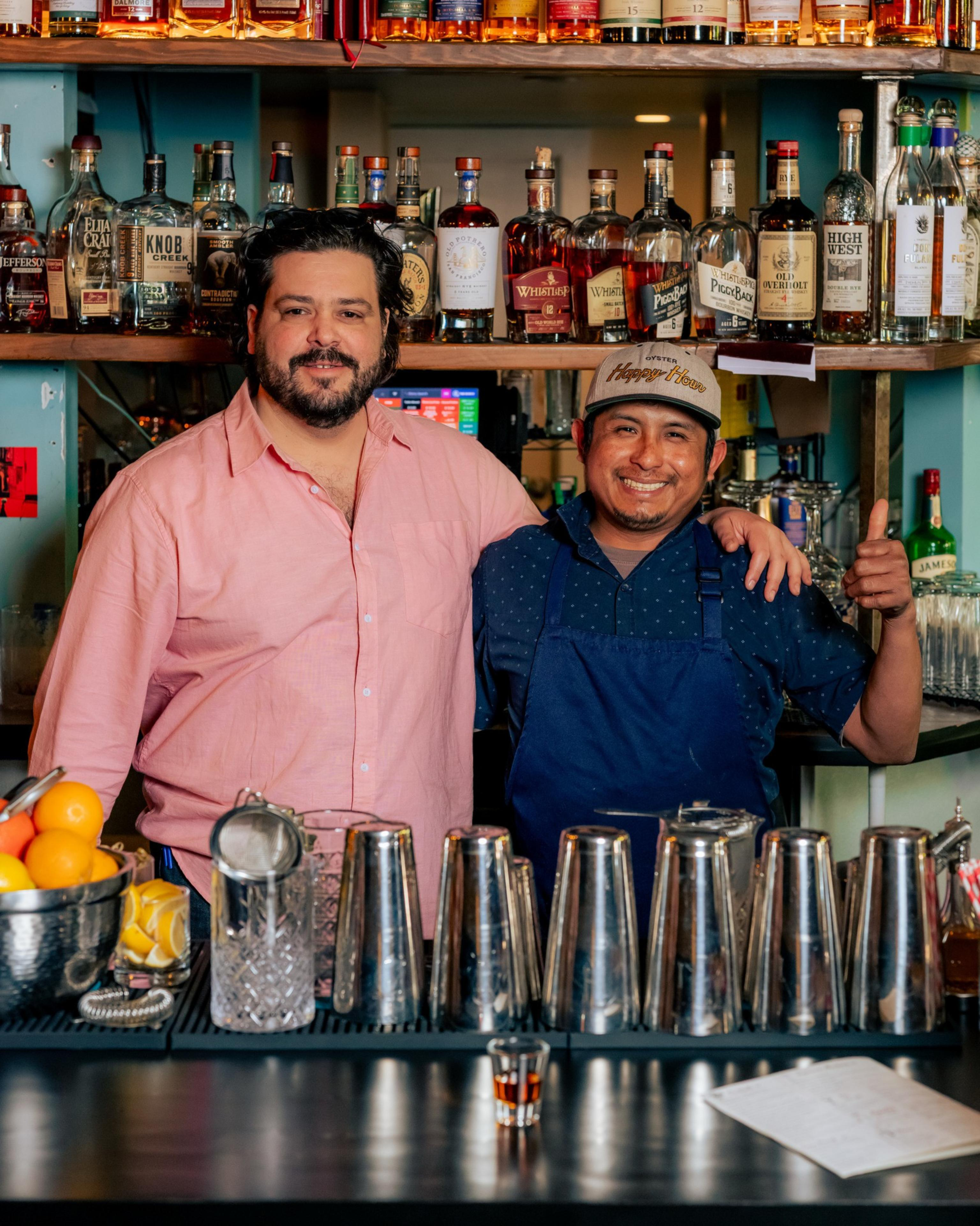 Two men stand behind a bar, smiling. One wears a pink shirt, and the other a blue apron and cap. Bottles line the shelves behind them, and shakers are on the counter.
