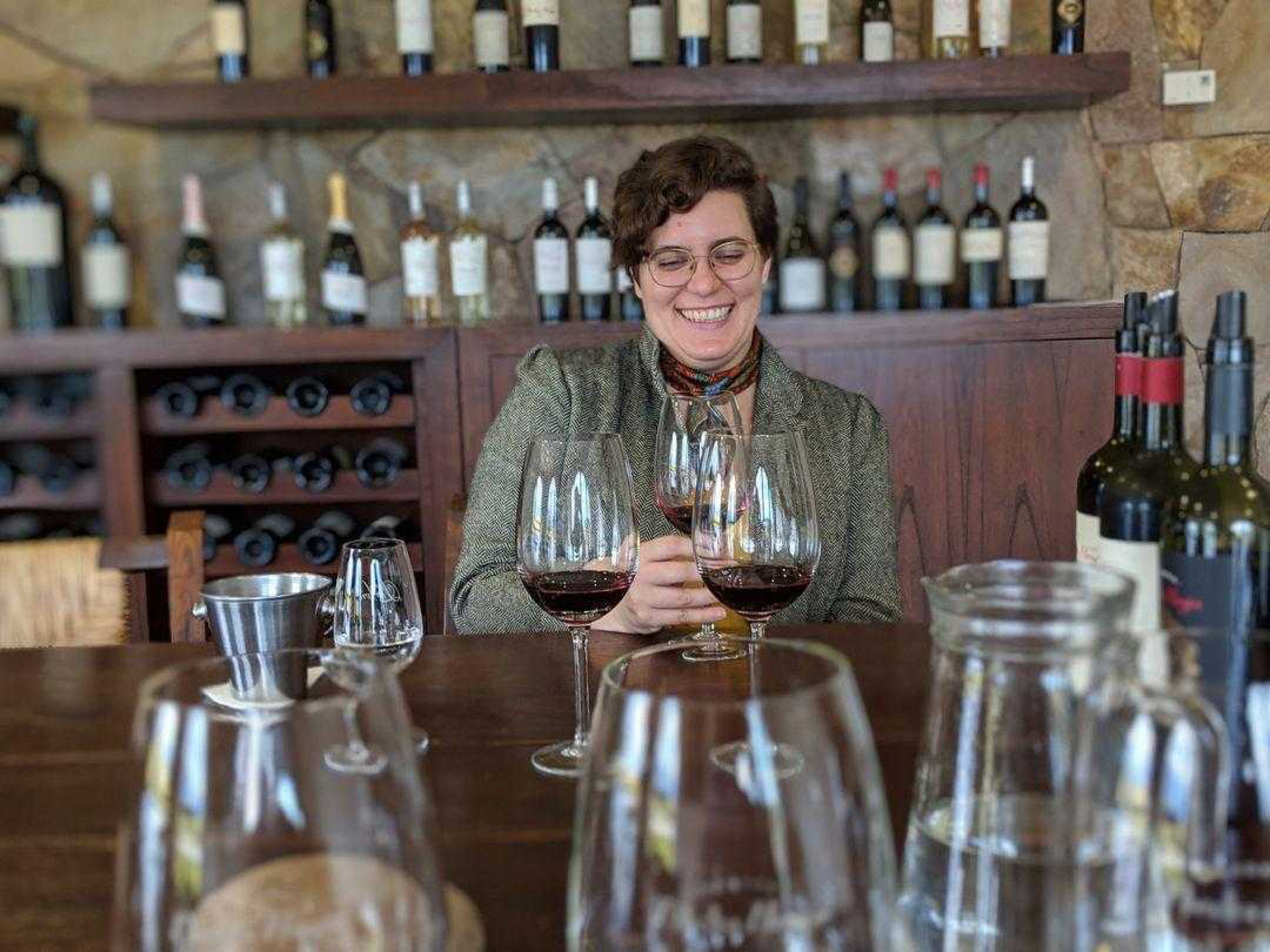 A smiling person with short curly hair and glasses sits at a table with several glasses of red wine and wine bottles behind them.