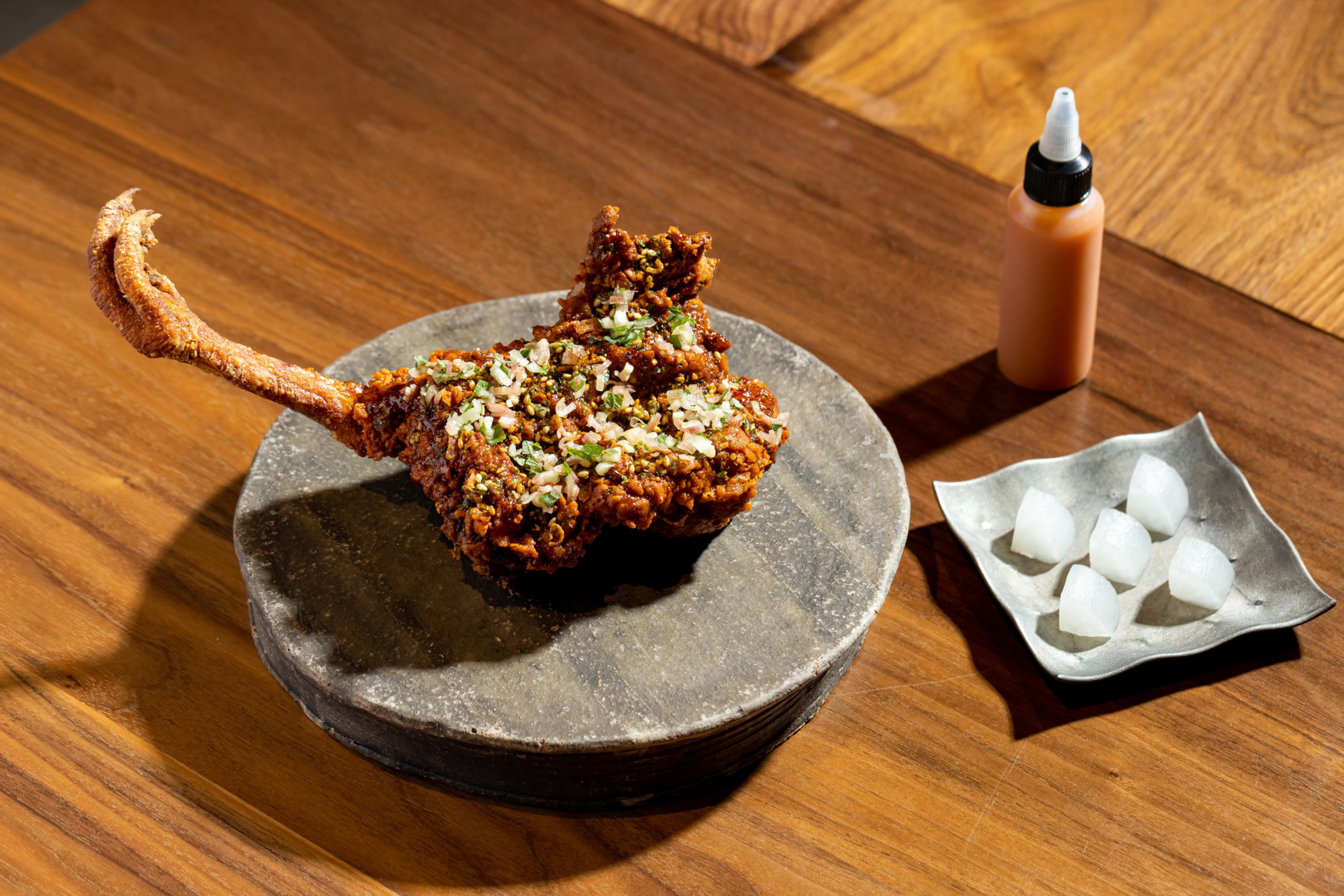 A large piece of crispy fried pork chop garnished with chopped herbs and onions rests on a round slate plate, next to a sauce bottle and a small dish of pickled onions.