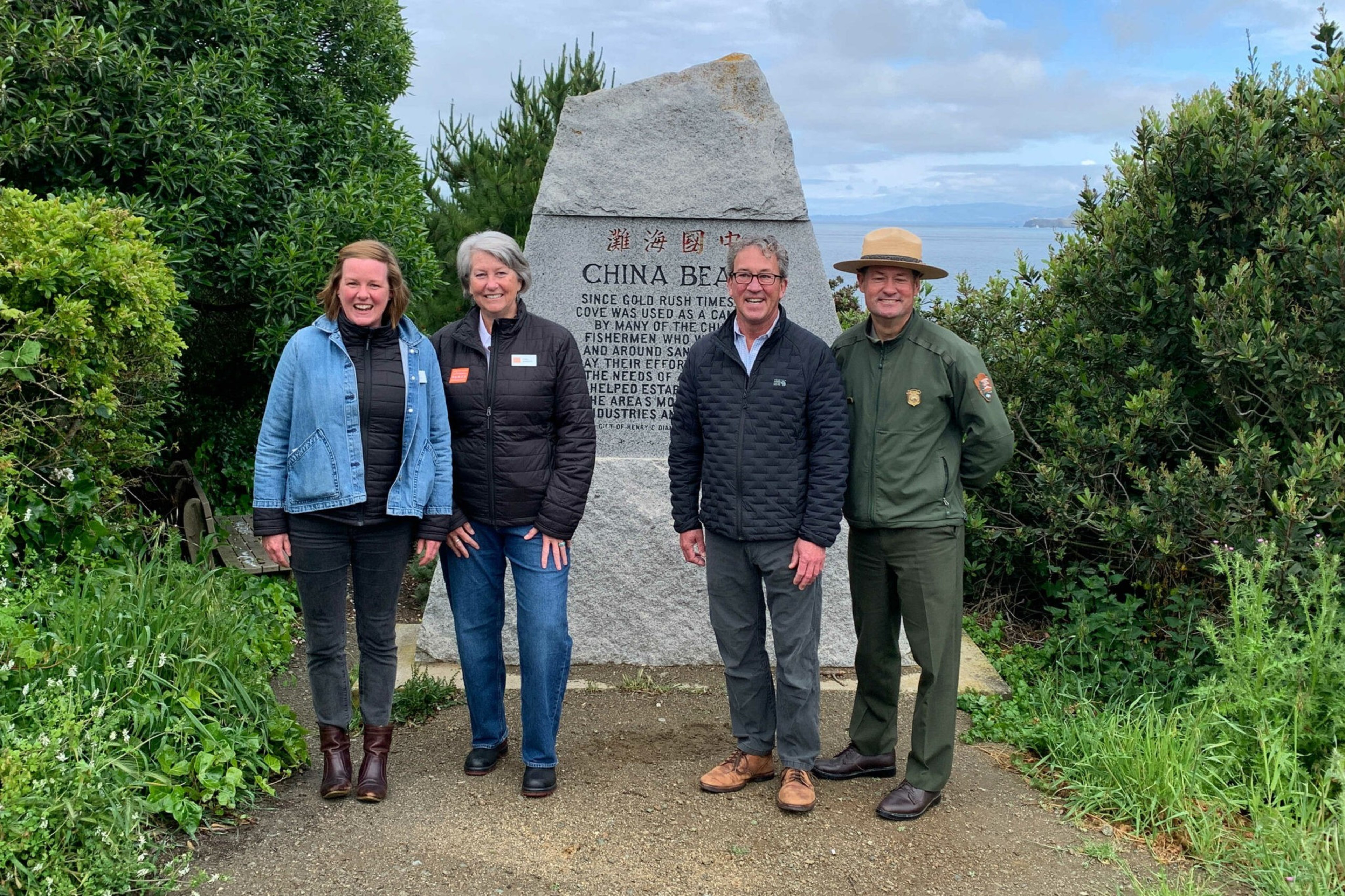 Four people stand smiling in front of a large stone monument with text about China Beach, surrounded by greenery and a partly cloudy sky.