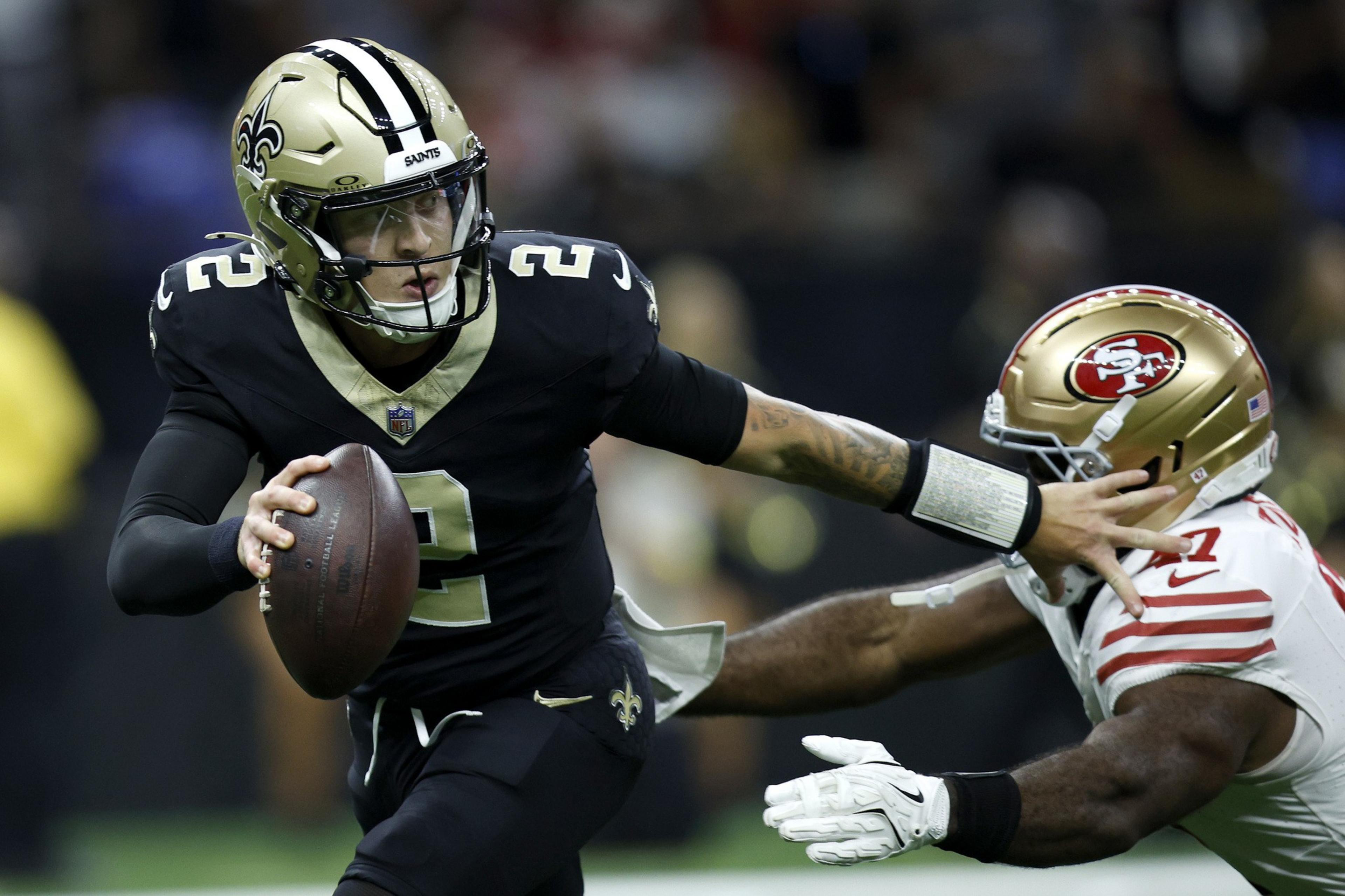 A football player in a black New Orleans Saints uniform fends off a defender from the San Francisco 49ers with an outstretched arm while holding the ball.