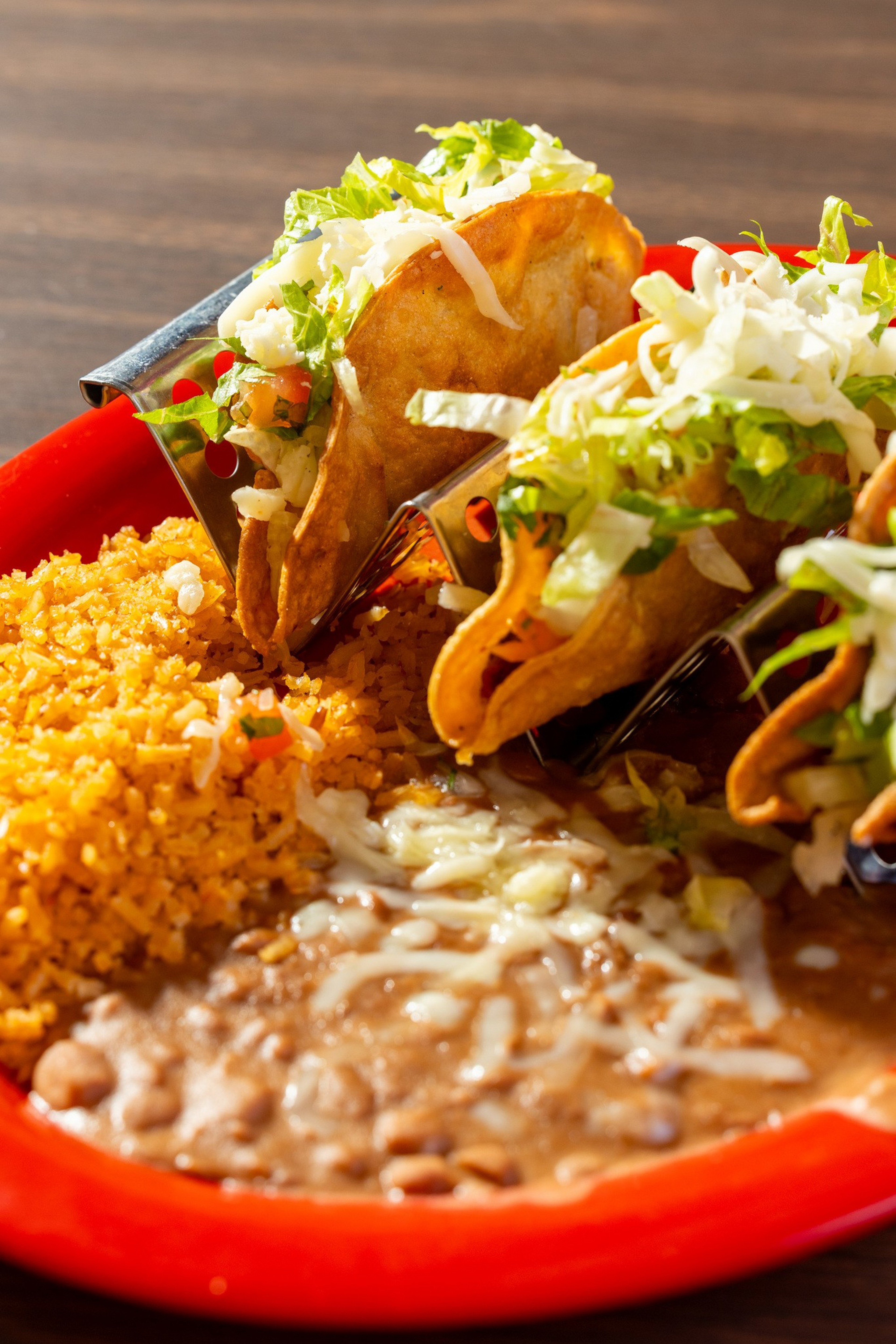 Three crispy tacos filled with lettuce, cheese, and other ingredients rest in a holder beside Mexican rice and refried beans topped with melted cheese.
