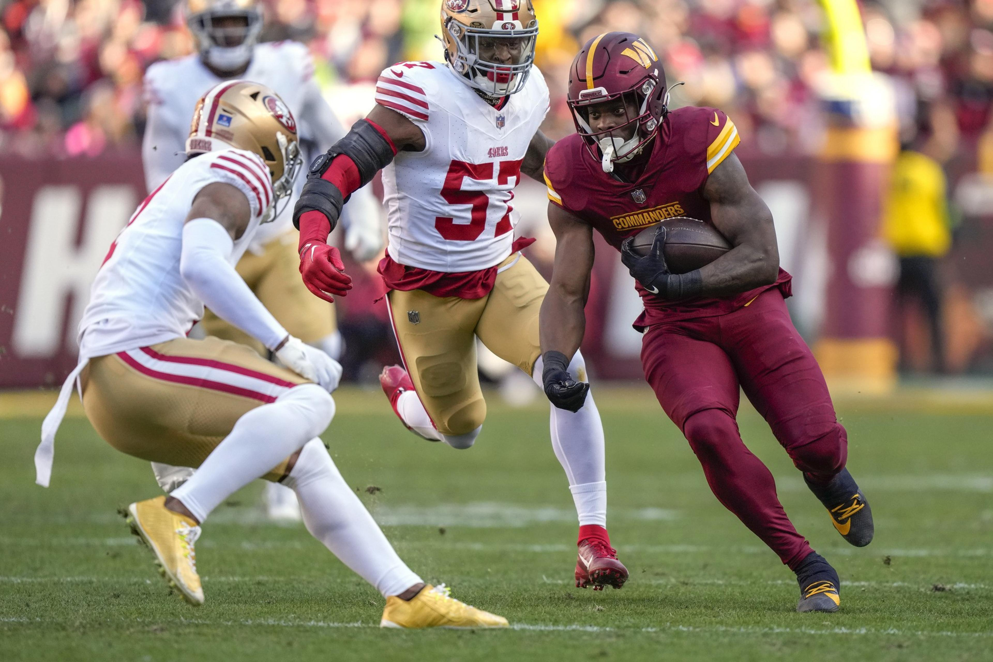 A football player in a red uniform runs with the ball while two players in white and gold uniforms try to tackle him on a grassy field.