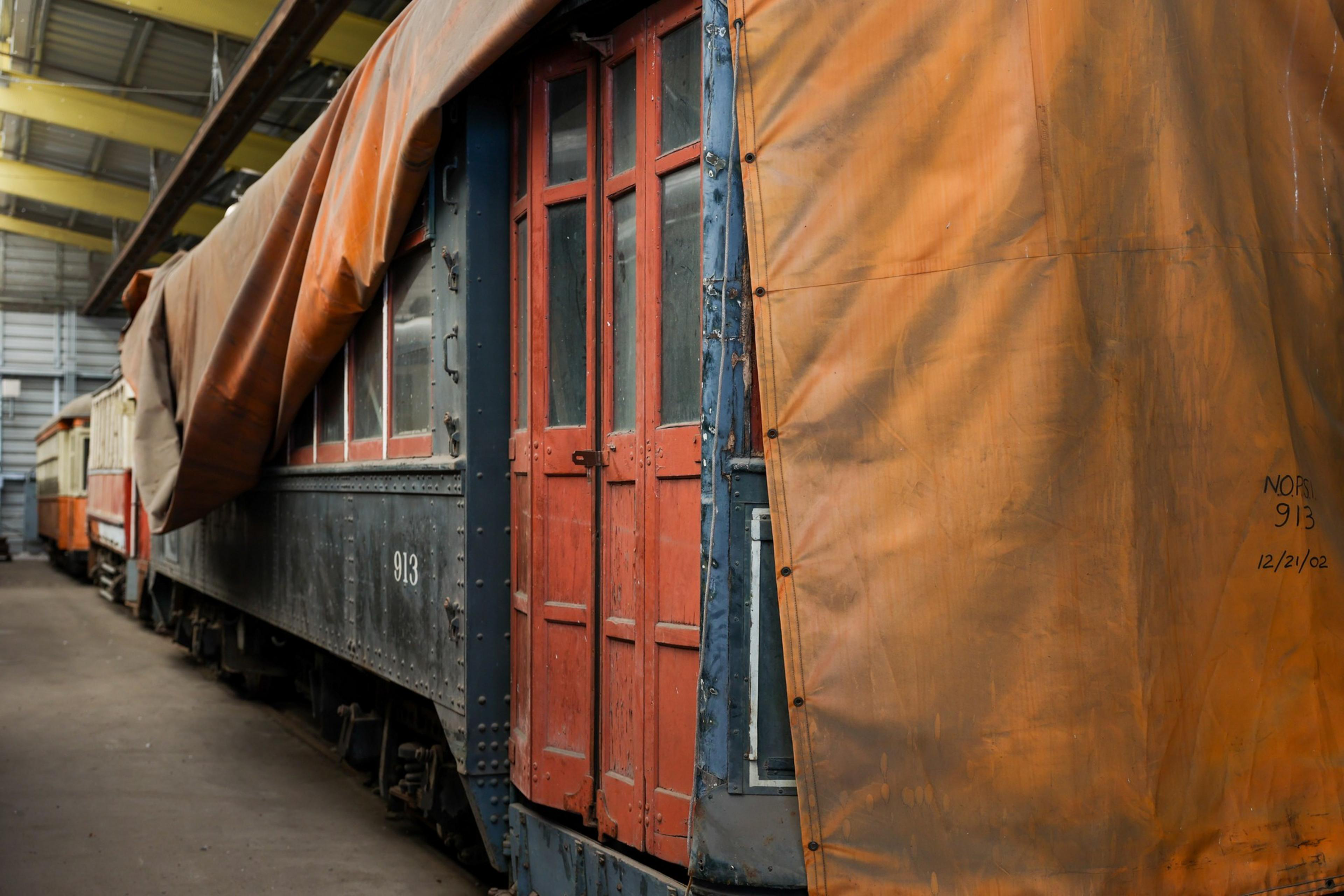 Several vintage train cars are parked inside a warehouse, with the closest one painted dark blue and featuring red doors partially covered by a large orange tarp.
