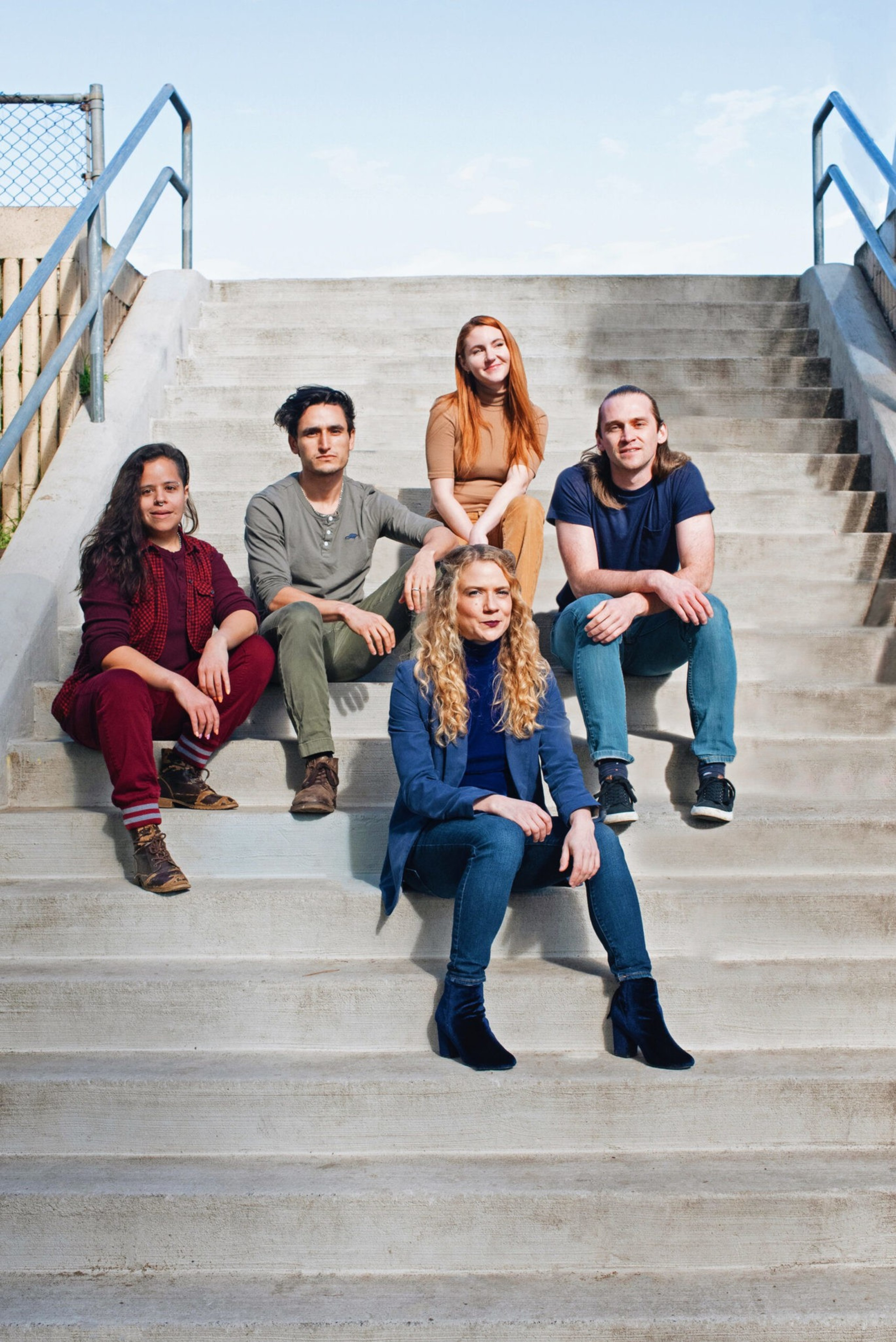 Five people sit on concrete stairs outdoors, posing casually with relaxed expressions and dressed in varied casual clothing.