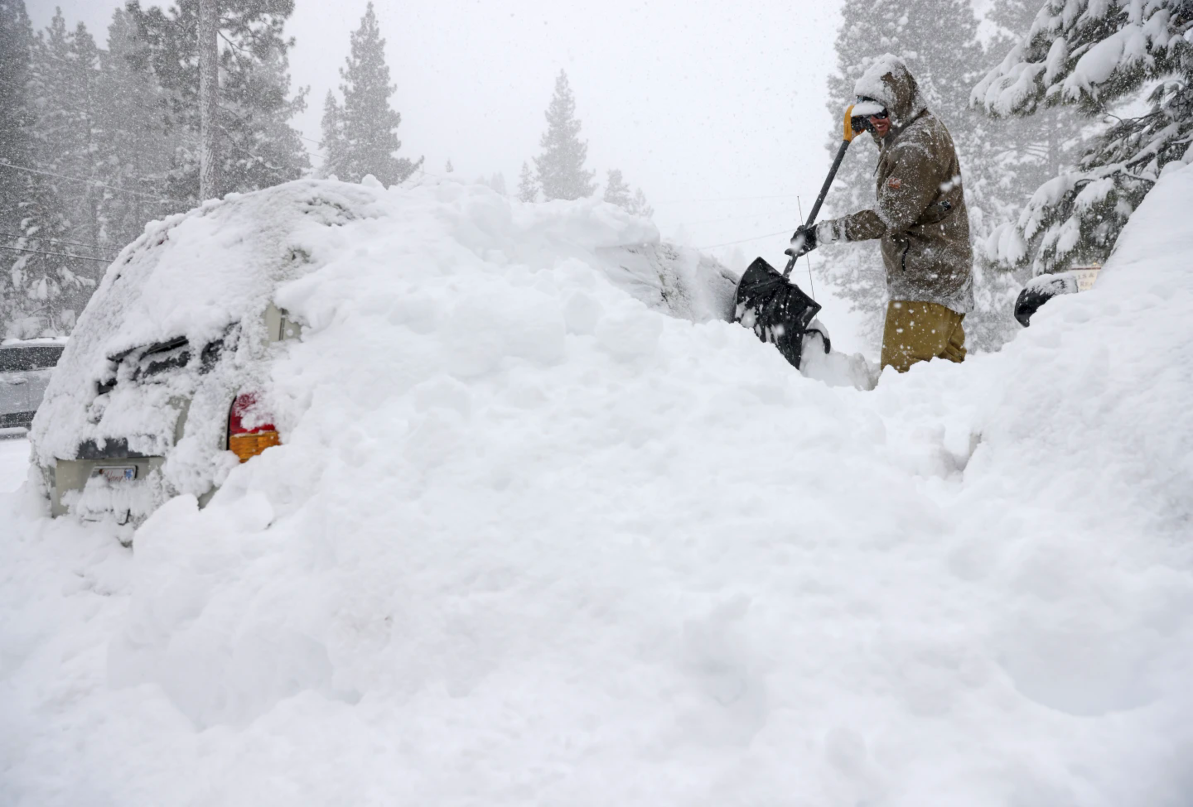 A person in winter gear shovels heavy snow engulfing a vehicle amidst a snowfall.