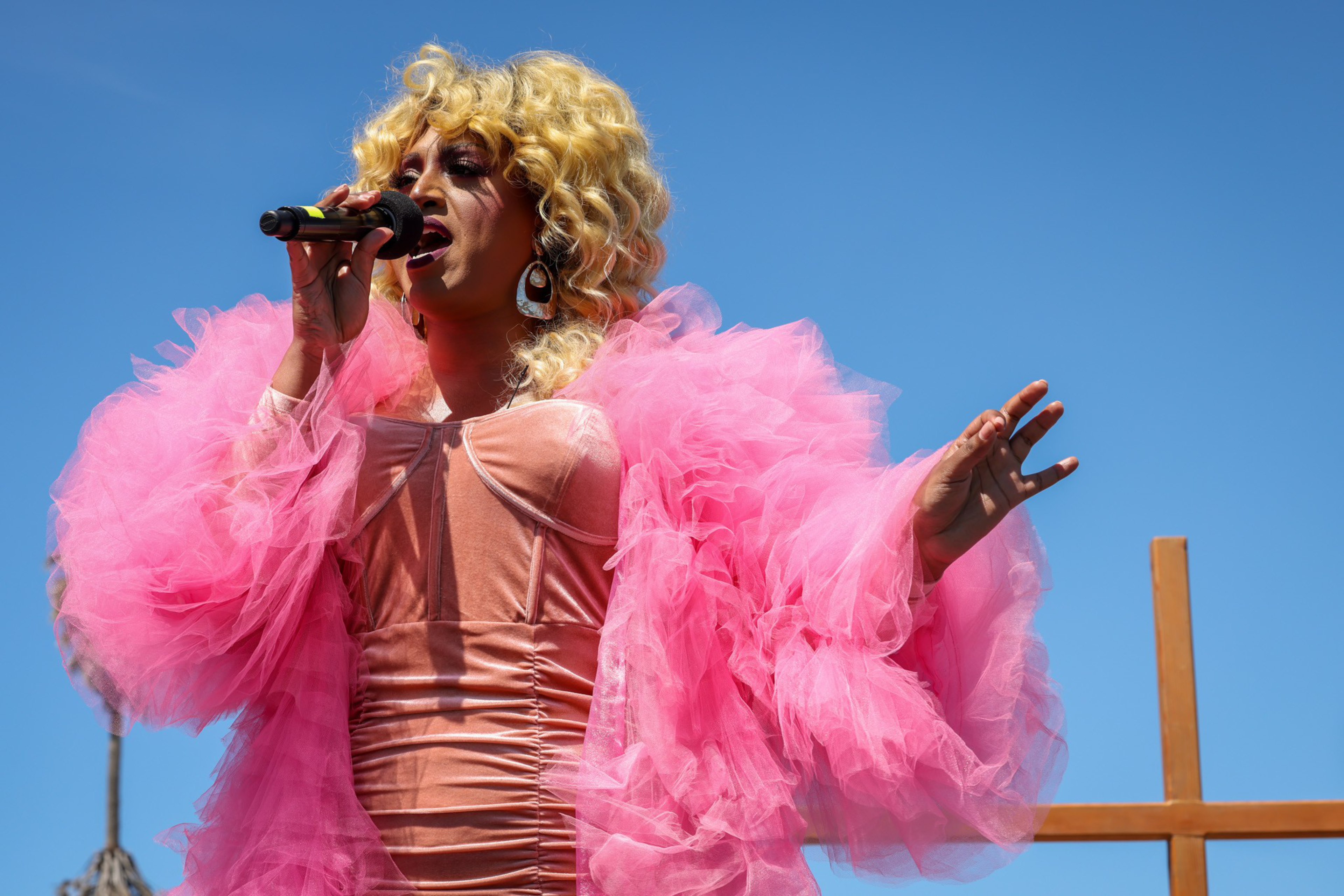 A performer with blonde curly hair sings into a microphone, wearing oversized pink tulle sleeves and a shiny fitted dress against a clear blue sky.
