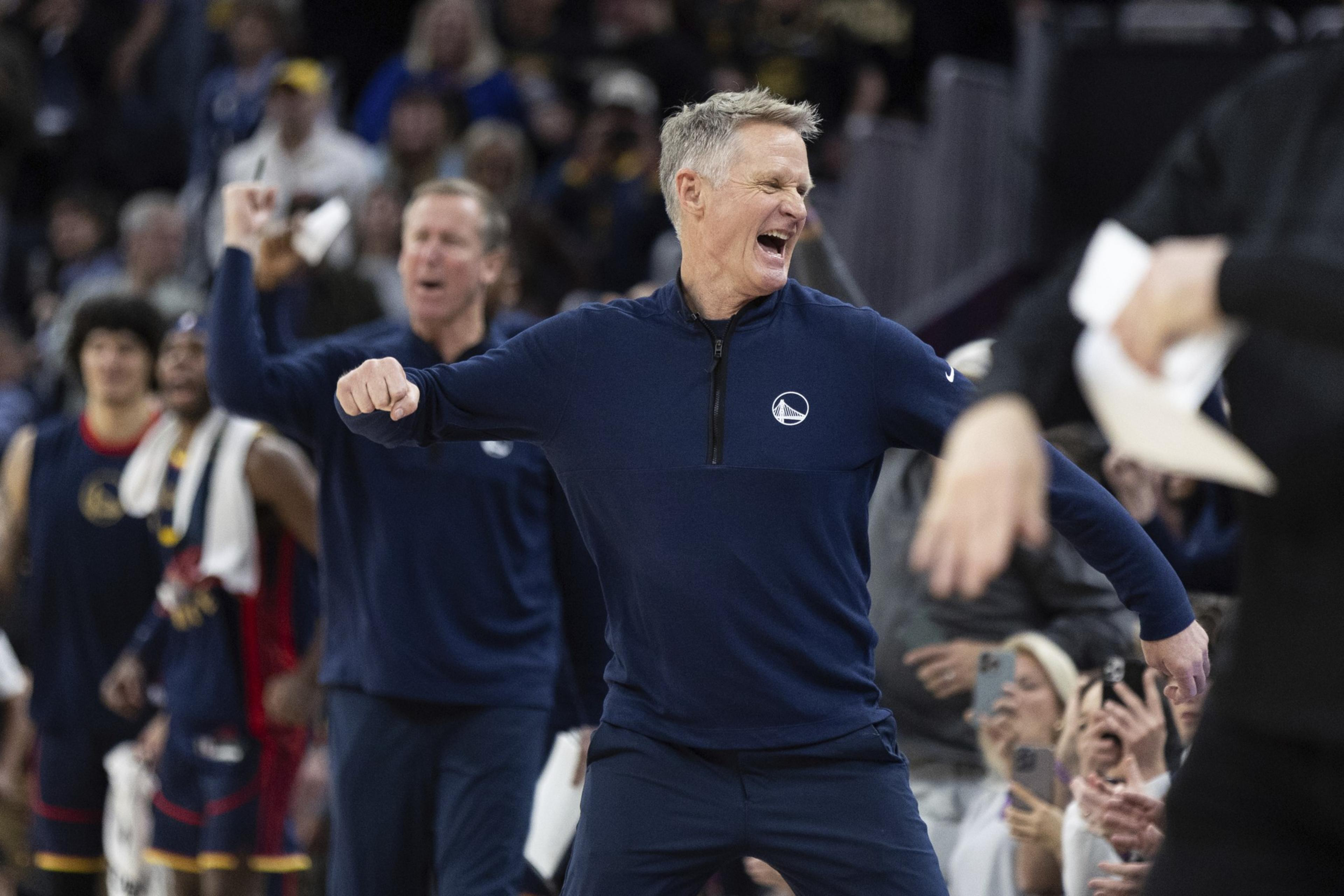 A man in a dark sports jacket is enthusiastically celebrating in a crowded basketball arena, surrounded by cheering fans and team members.