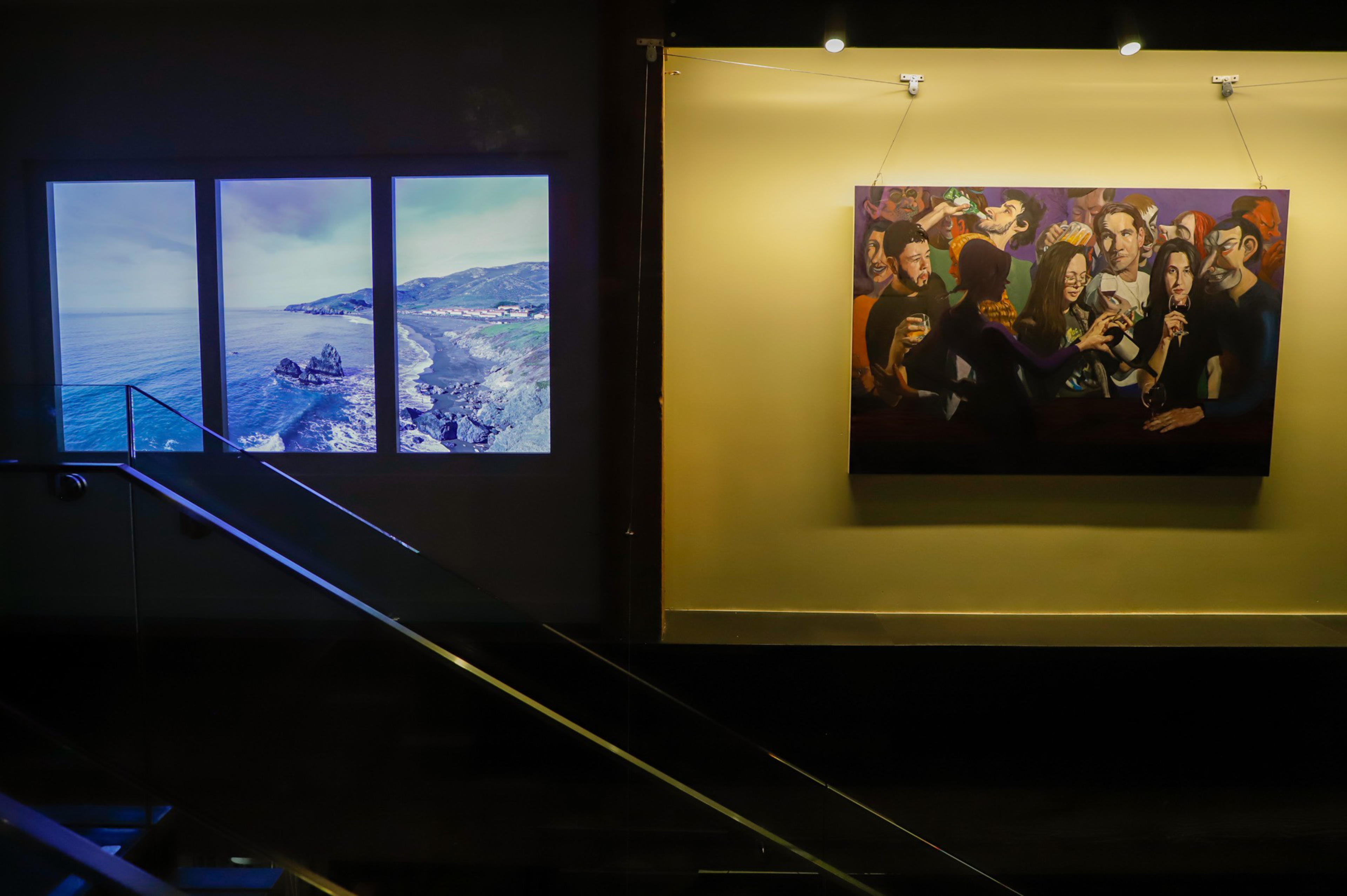A dark room features a three-panel window showing a coastal scene on the left and a colorful painting of people socializing on the right wall.
