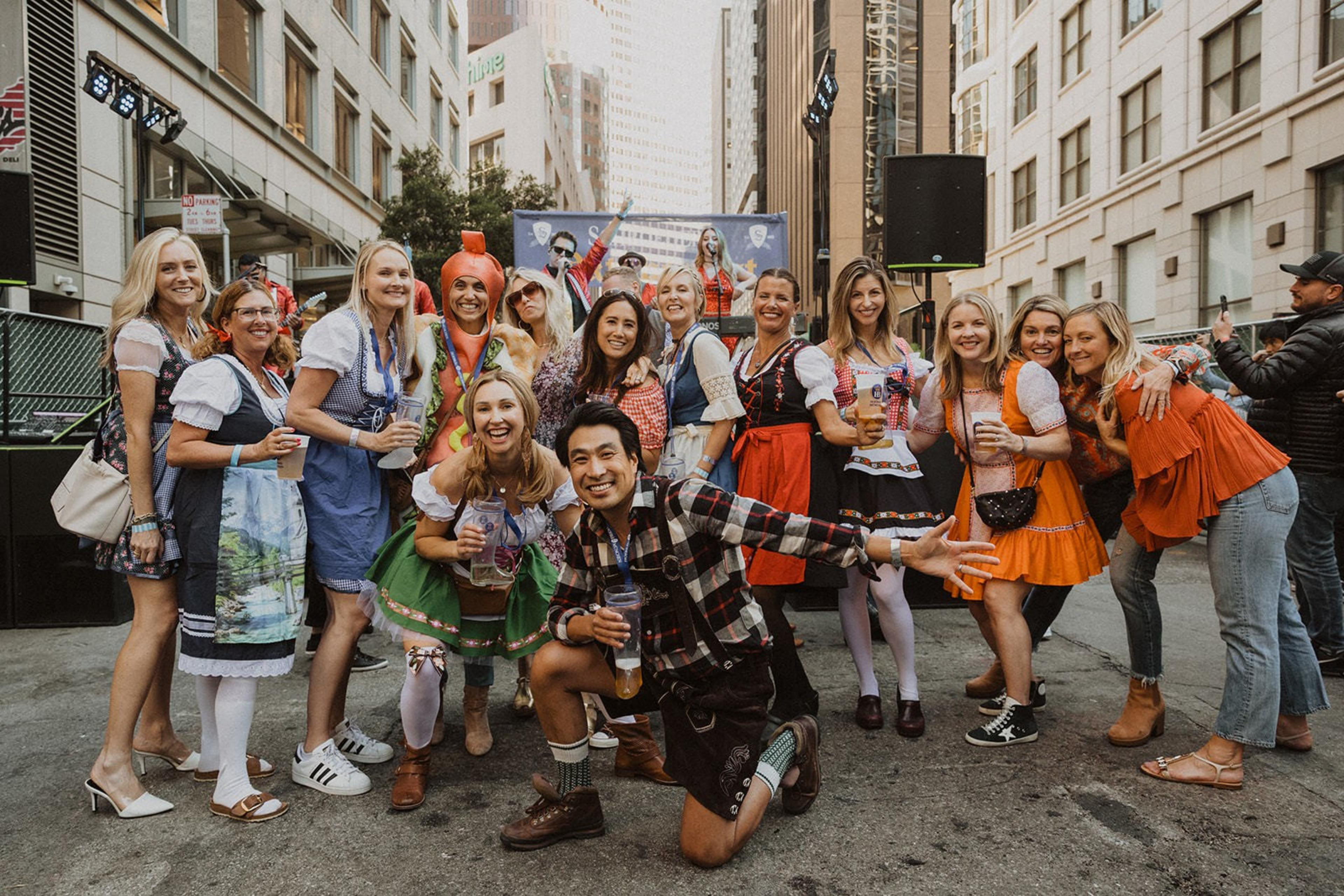 A vibrant group of people dressed in traditional Bavarian outfits and costumes pose joyfully in a city street, enjoying a festive event with tall buildings in the background.