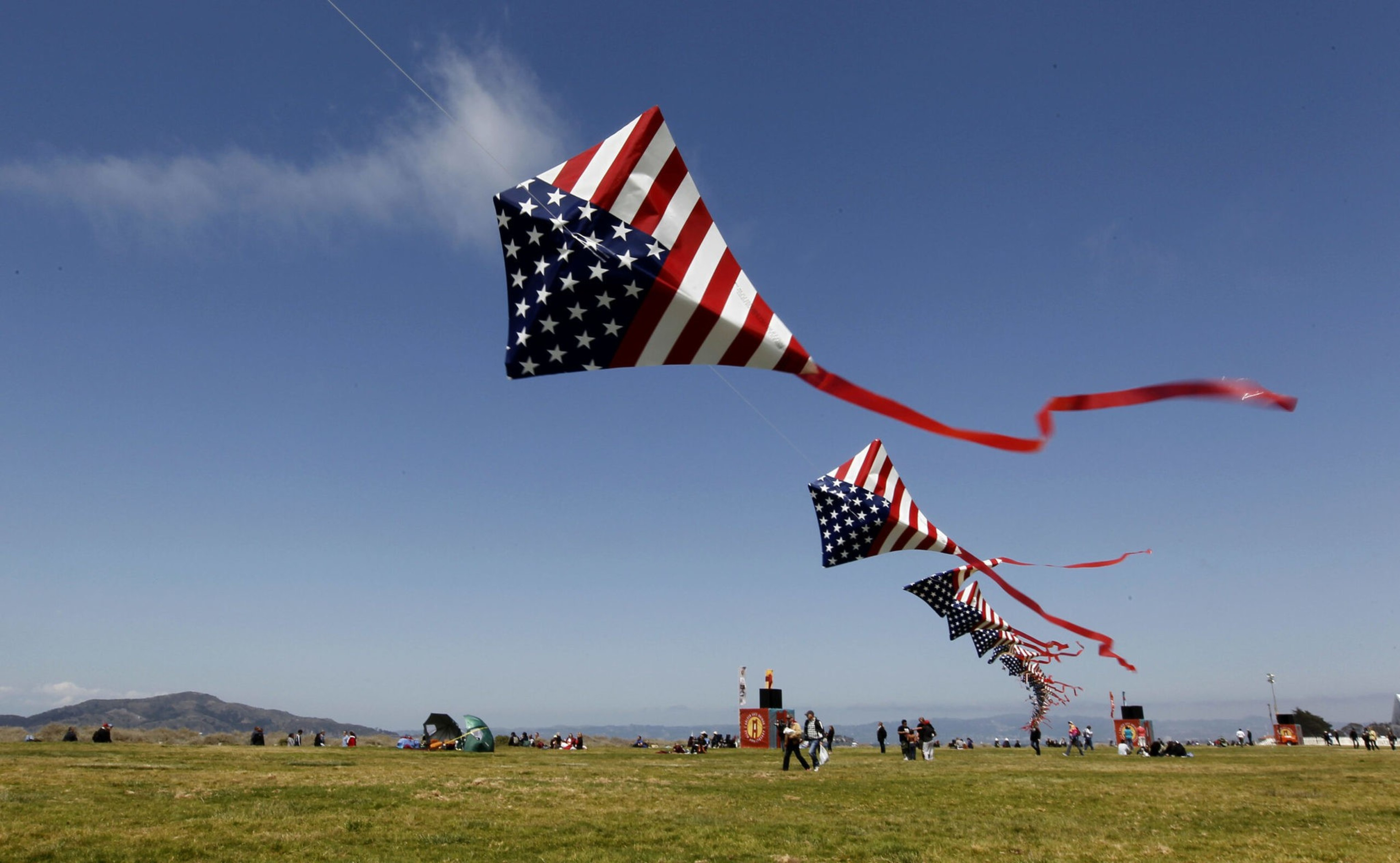 Several large kites with the American flag pattern fly in a line above a grassy field with people walking and sitting under a clear blue sky.