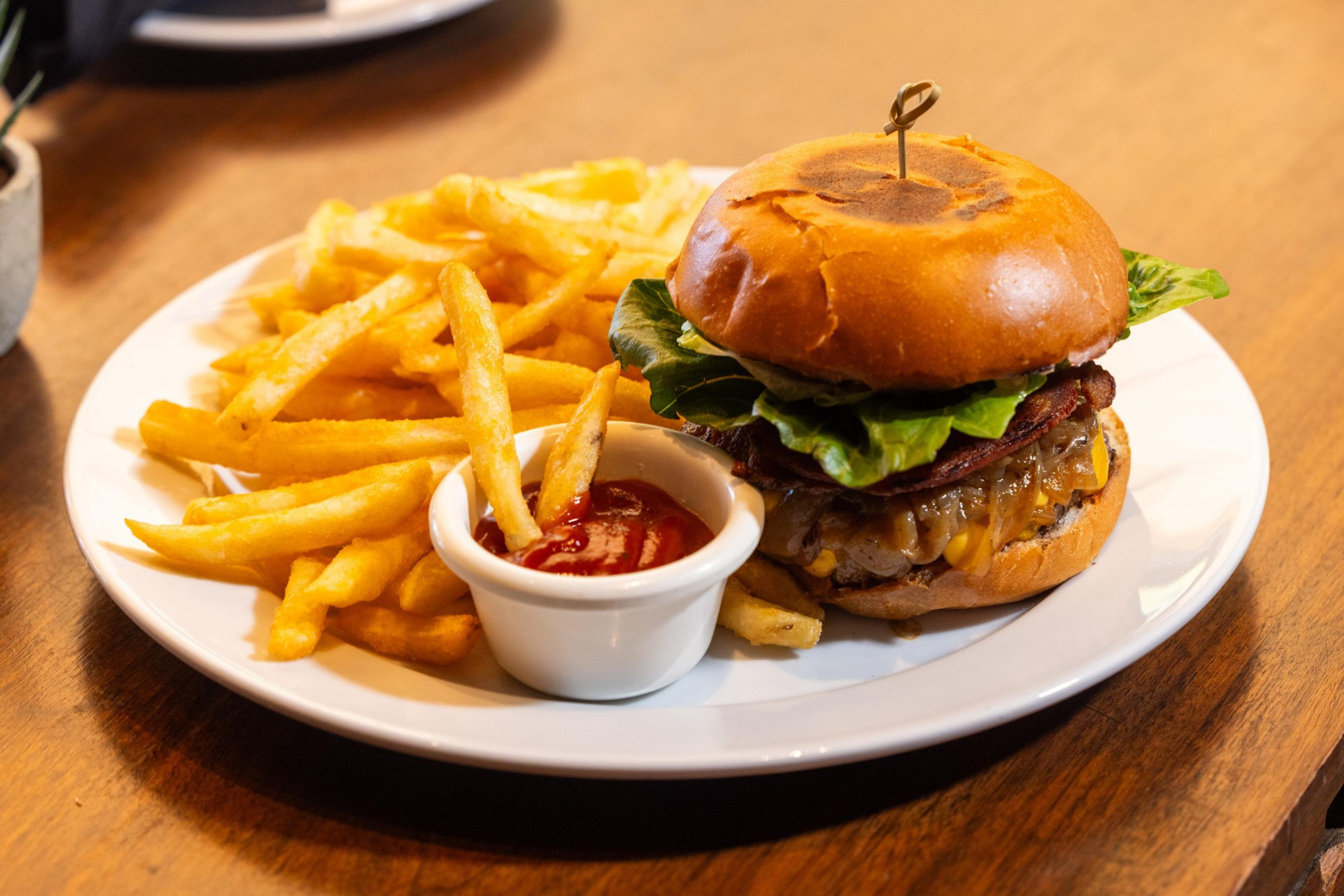 A cheeseburger with bacon and lettuce sits on a white plate next to golden French fries and a small cup of ketchup.