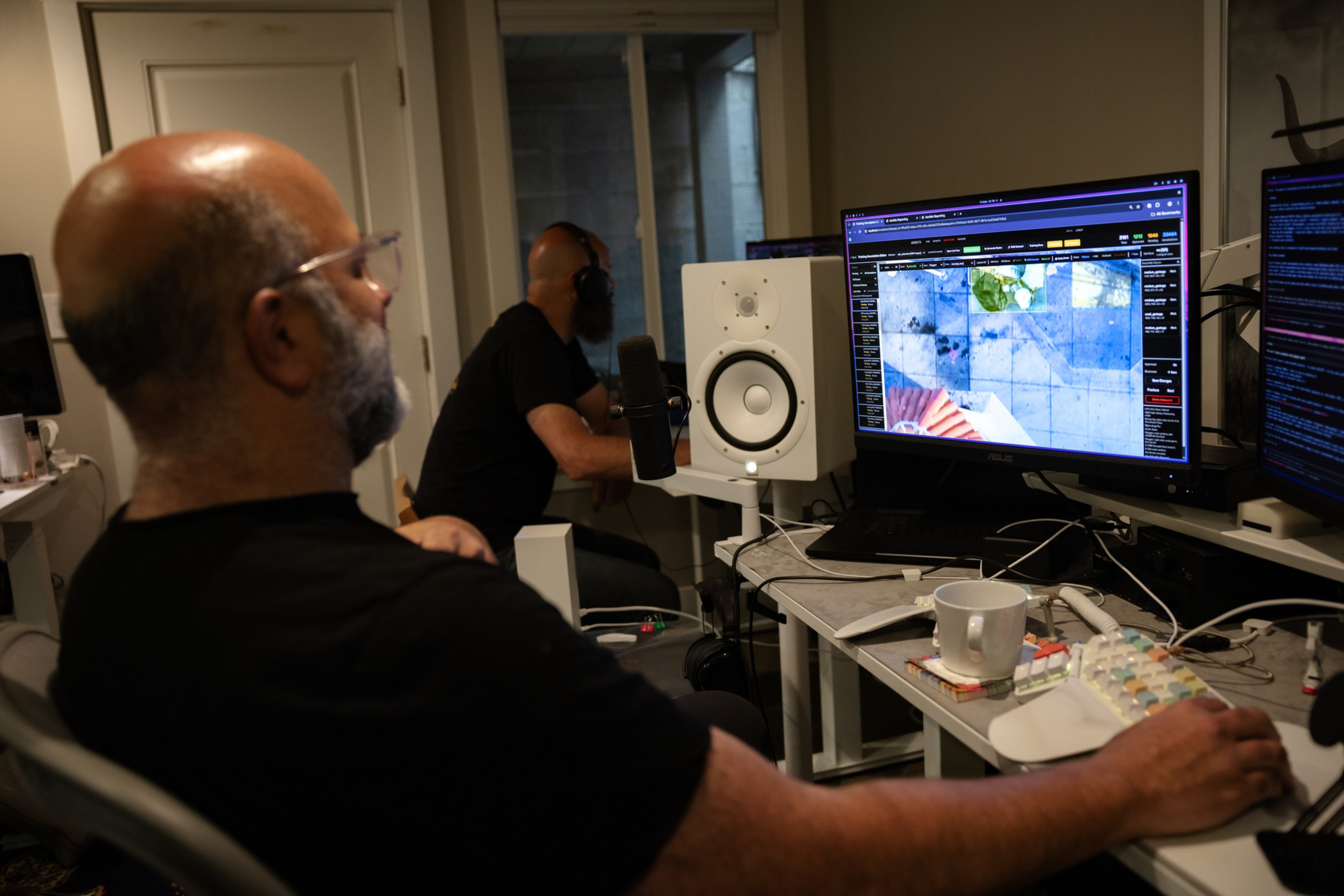 A man with glasses and a beard works at a computer displaying satellite or aerial imagery, with another man using headphones in the background.