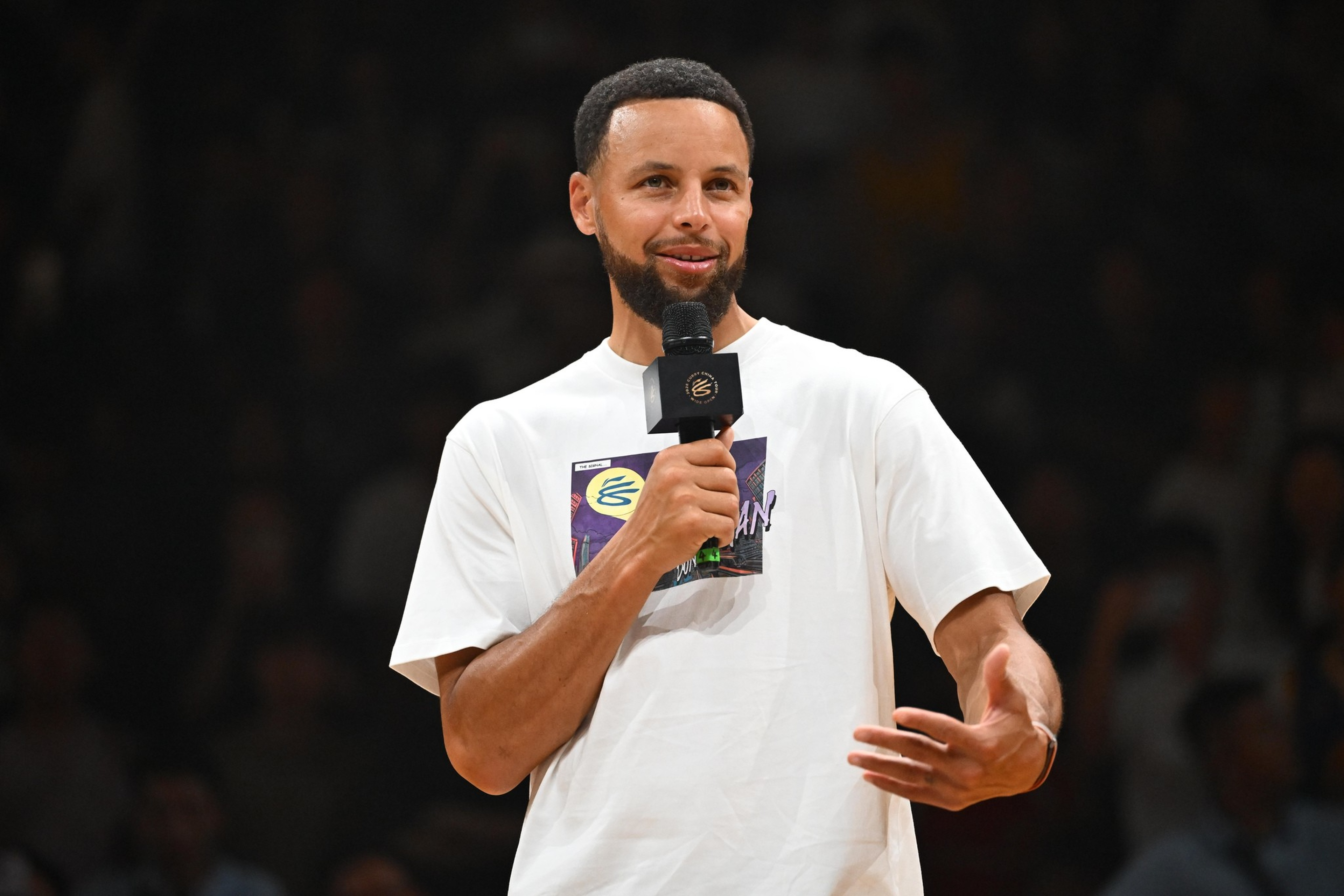 A man with a beard speaks into a microphone, wearing a white graphic T-shirt, gesturing with one hand in front of a dark blurred background.