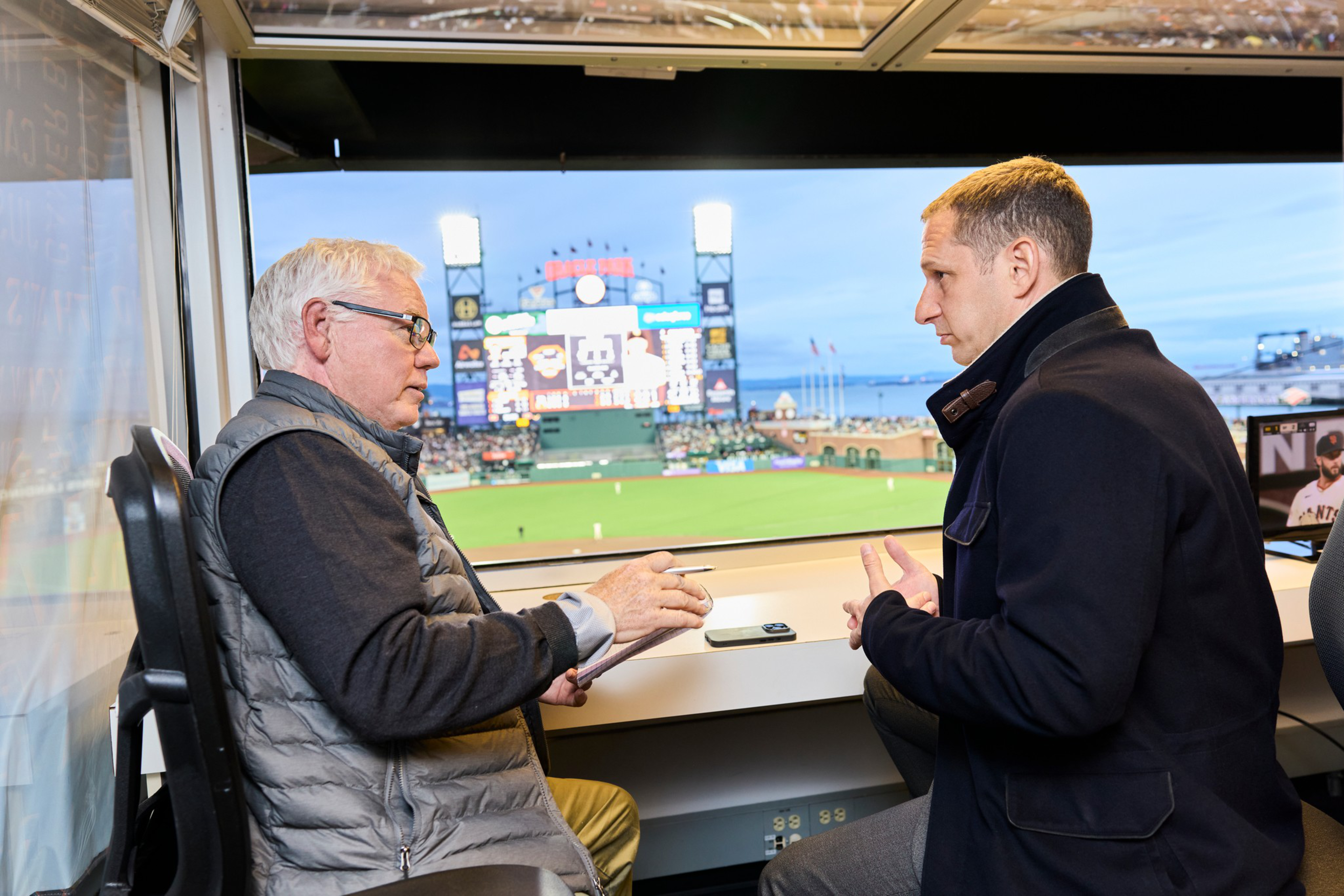 Two men are sitting and talking in a press box overlooking a baseball field during a game at dusk.