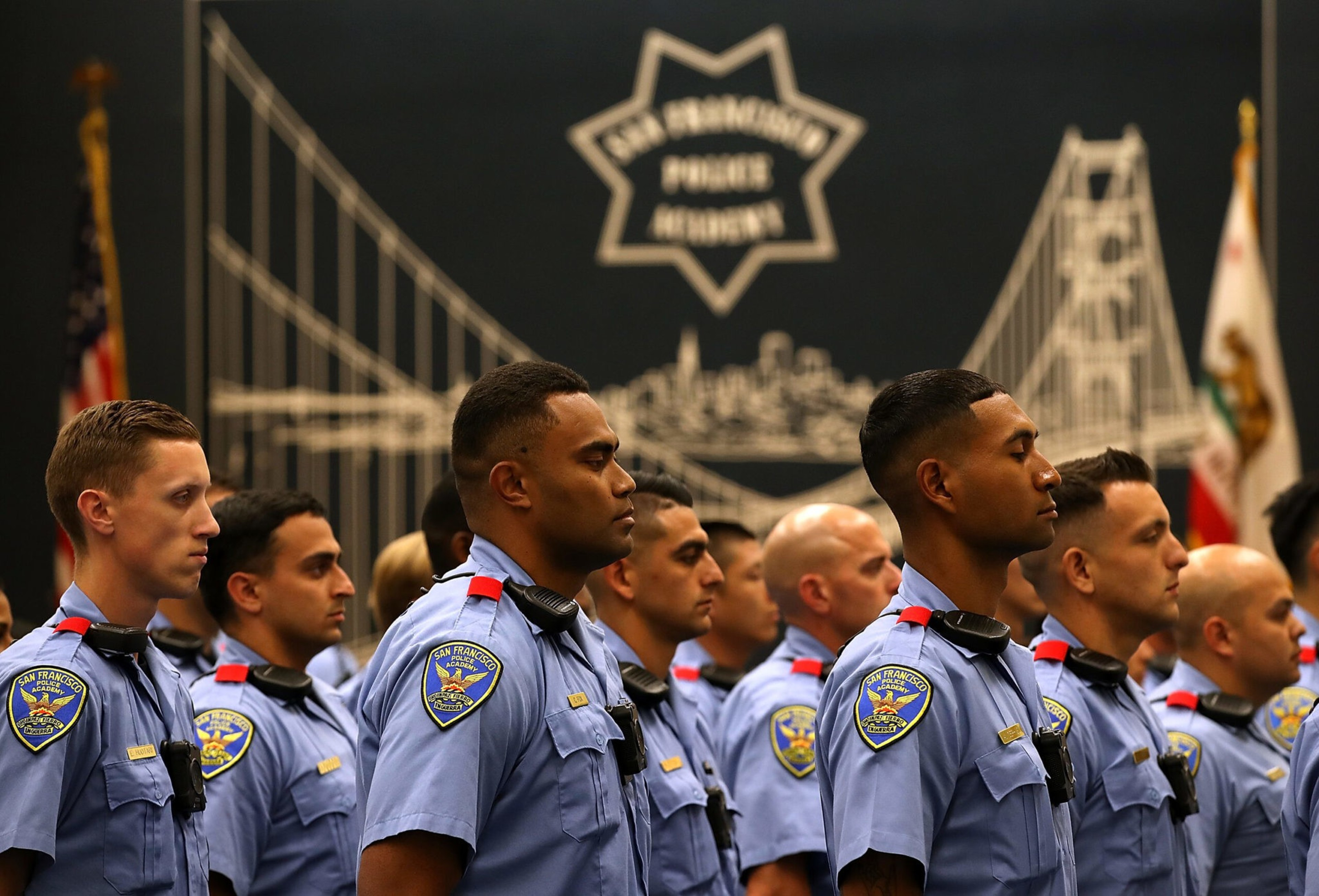 A row of police cadets in blue uniforms stand attentively in front of a backdrop featuring the Golden Gate Bridge, the text &quot;San Francisco Police Academy,&quot; and two flags.