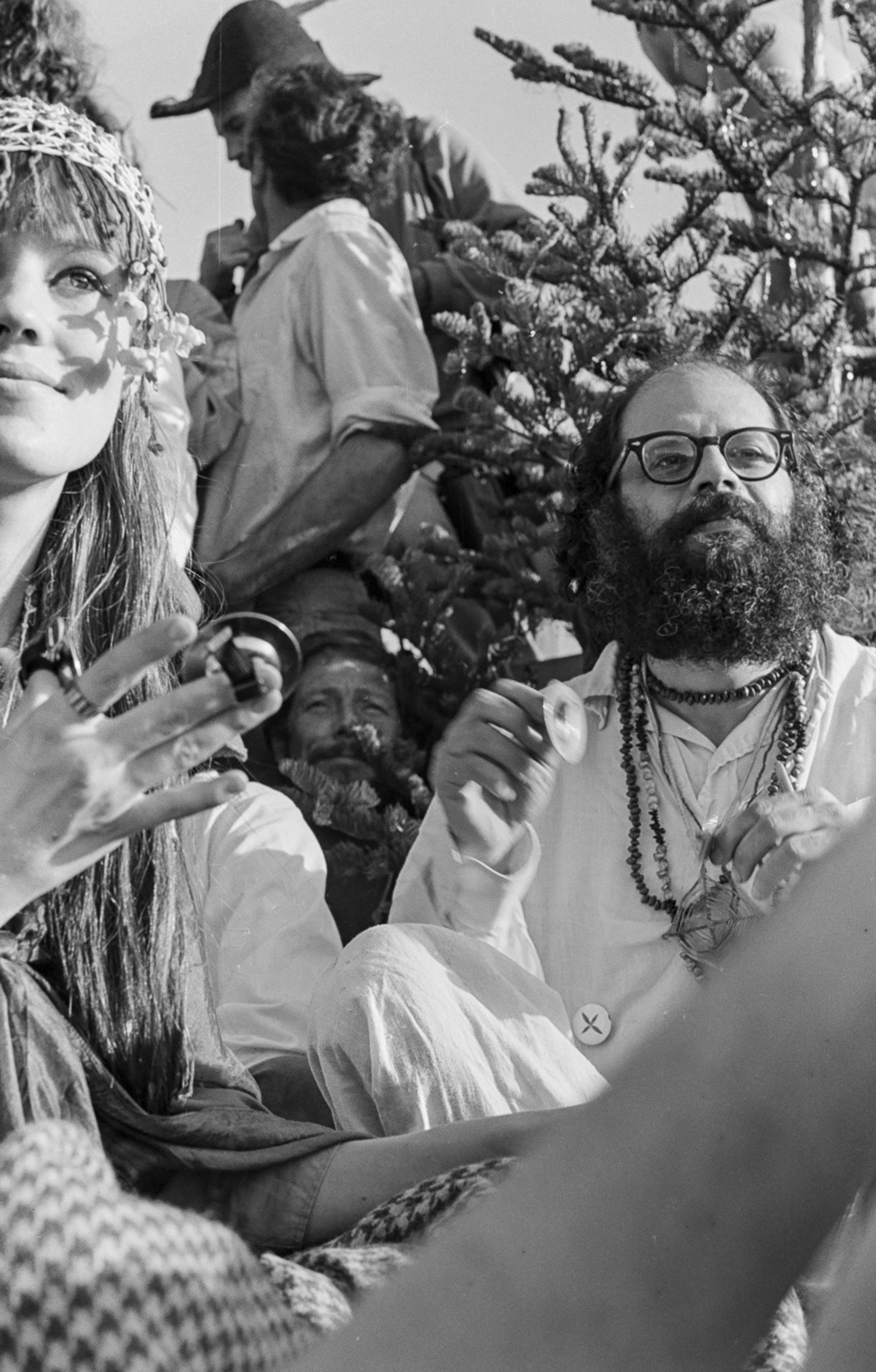 A black-and-white photo shows a group of people outdoors. A woman with a flower headband smiles on the left, and a man with glasses and a beard holds an object on the right.