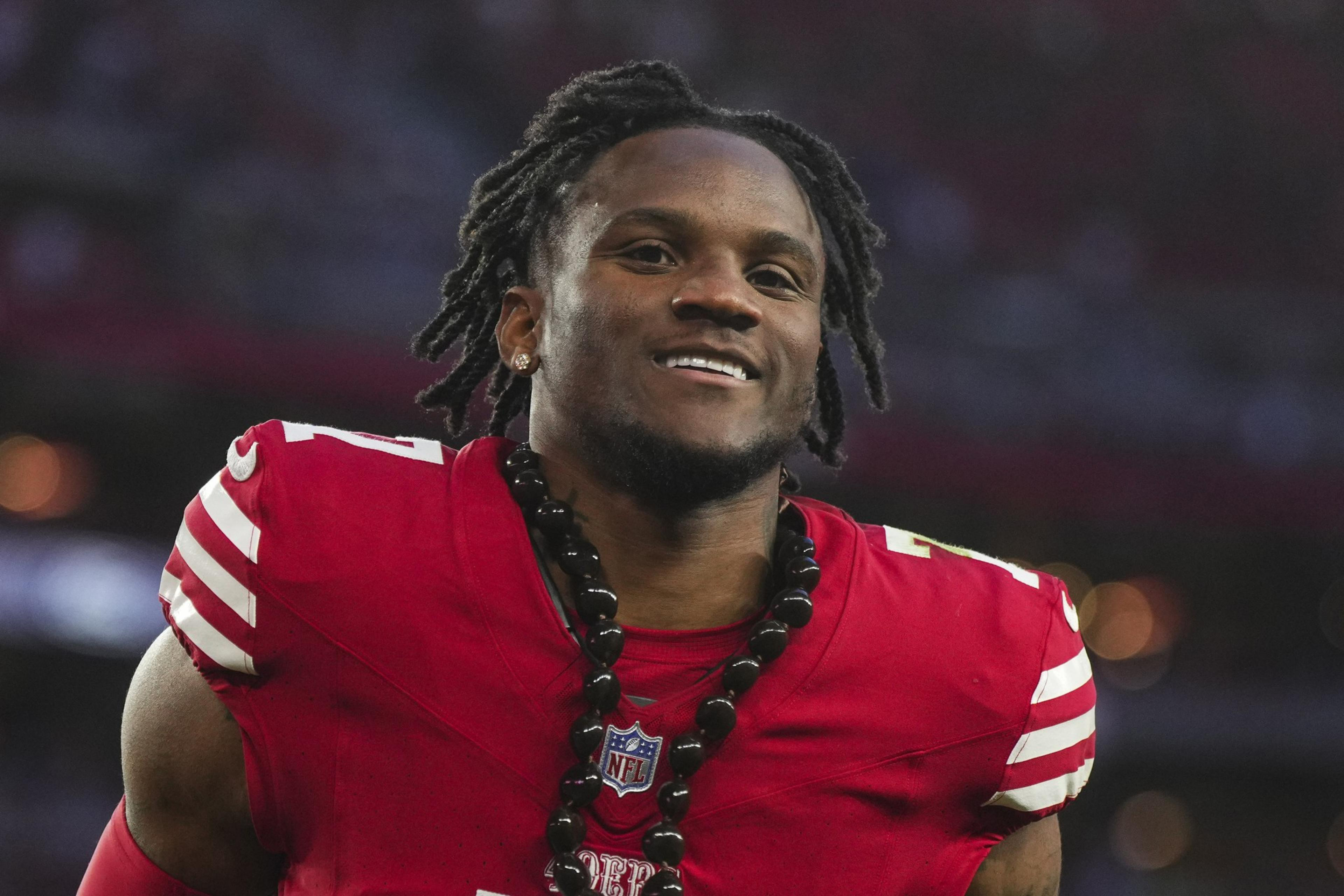 A smiling football player with dreadlocks in a red jersey adorned with a large bead necklace.