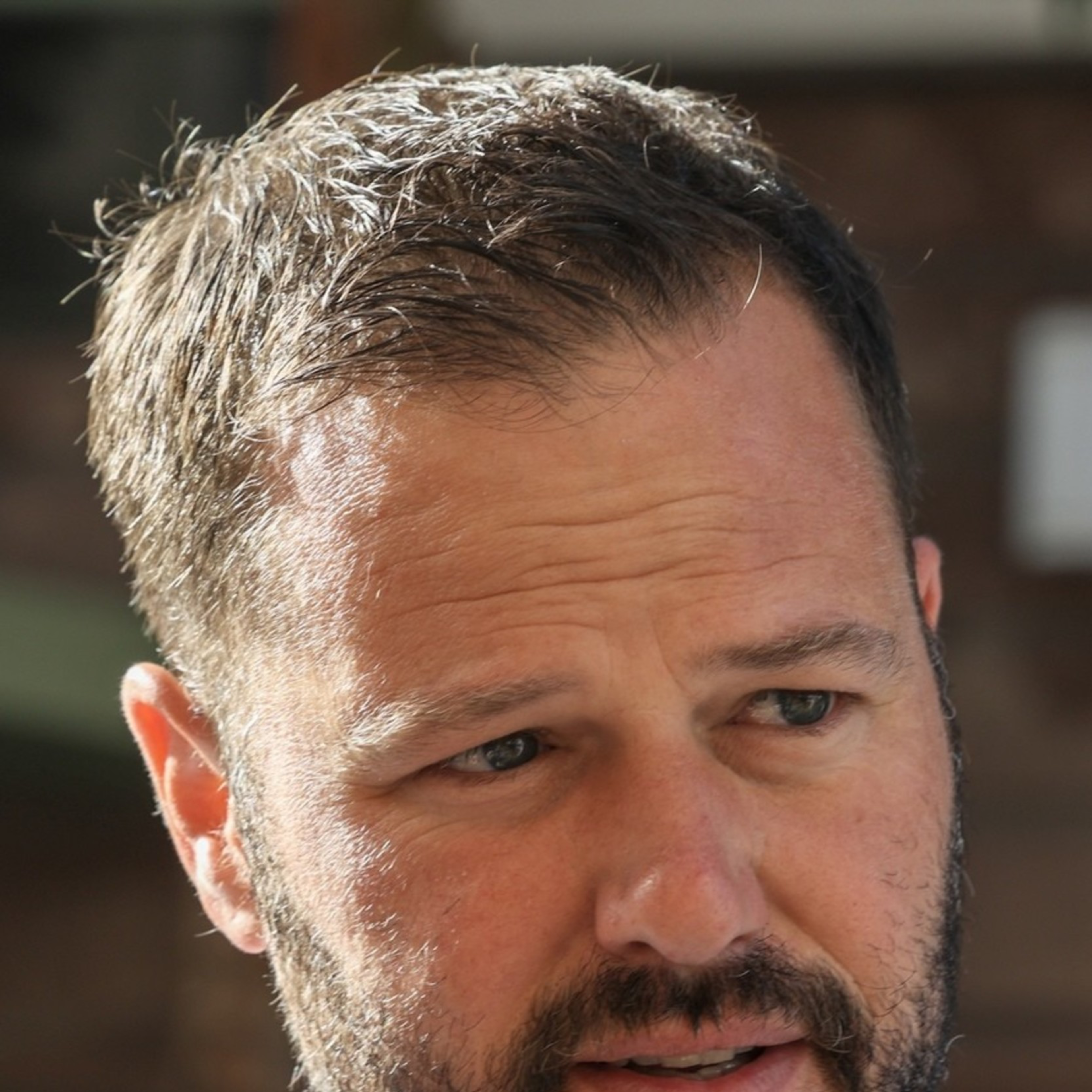 A man with short brown hair and a beard is wearing a white dress shirt and a striped tie. He is outdoors, set against a blurred background of a building facade.