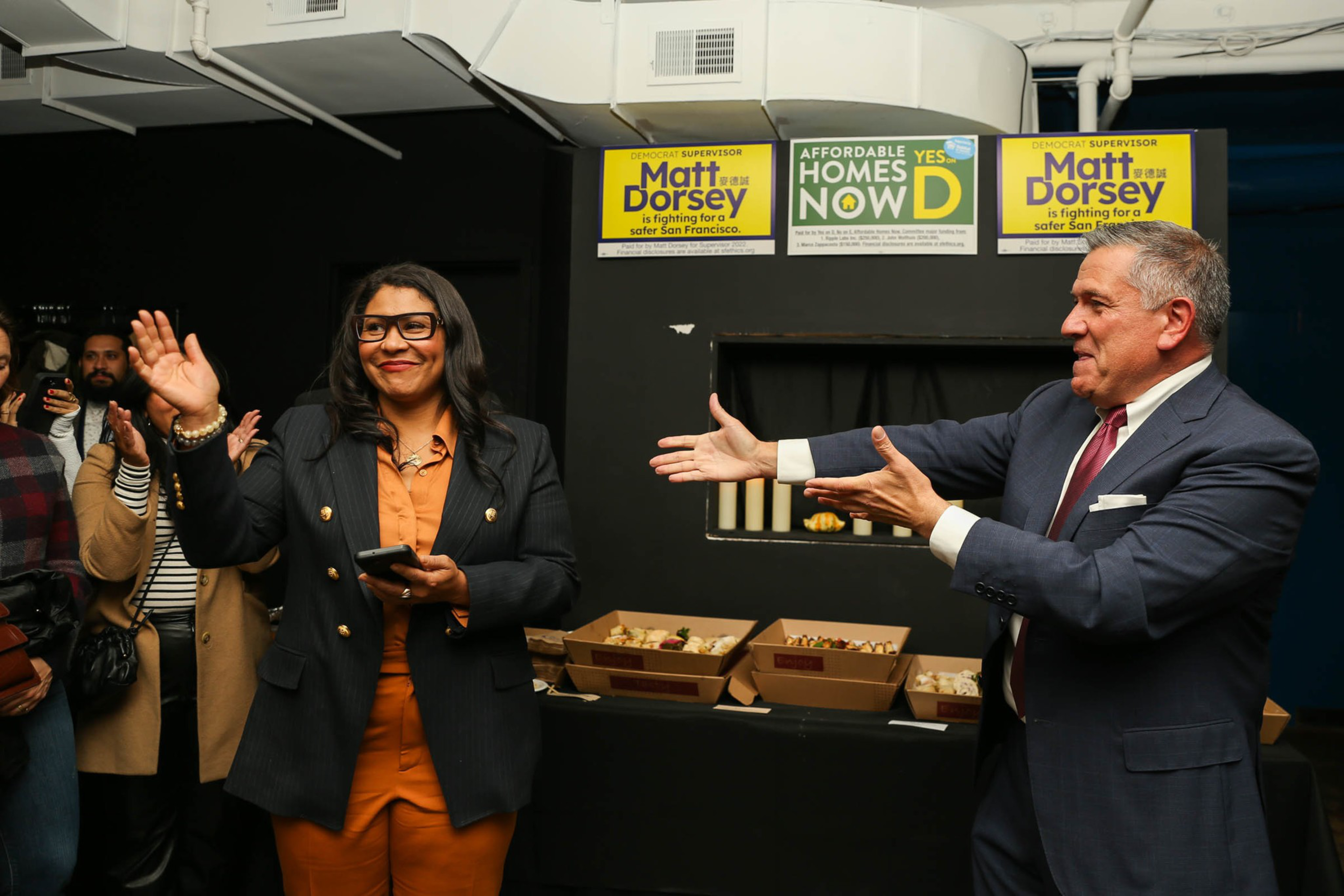 A woman in an orange outfit and black blazer waves while a man in a suit gestures toward her, with people clapping in the background and campaign signs above.