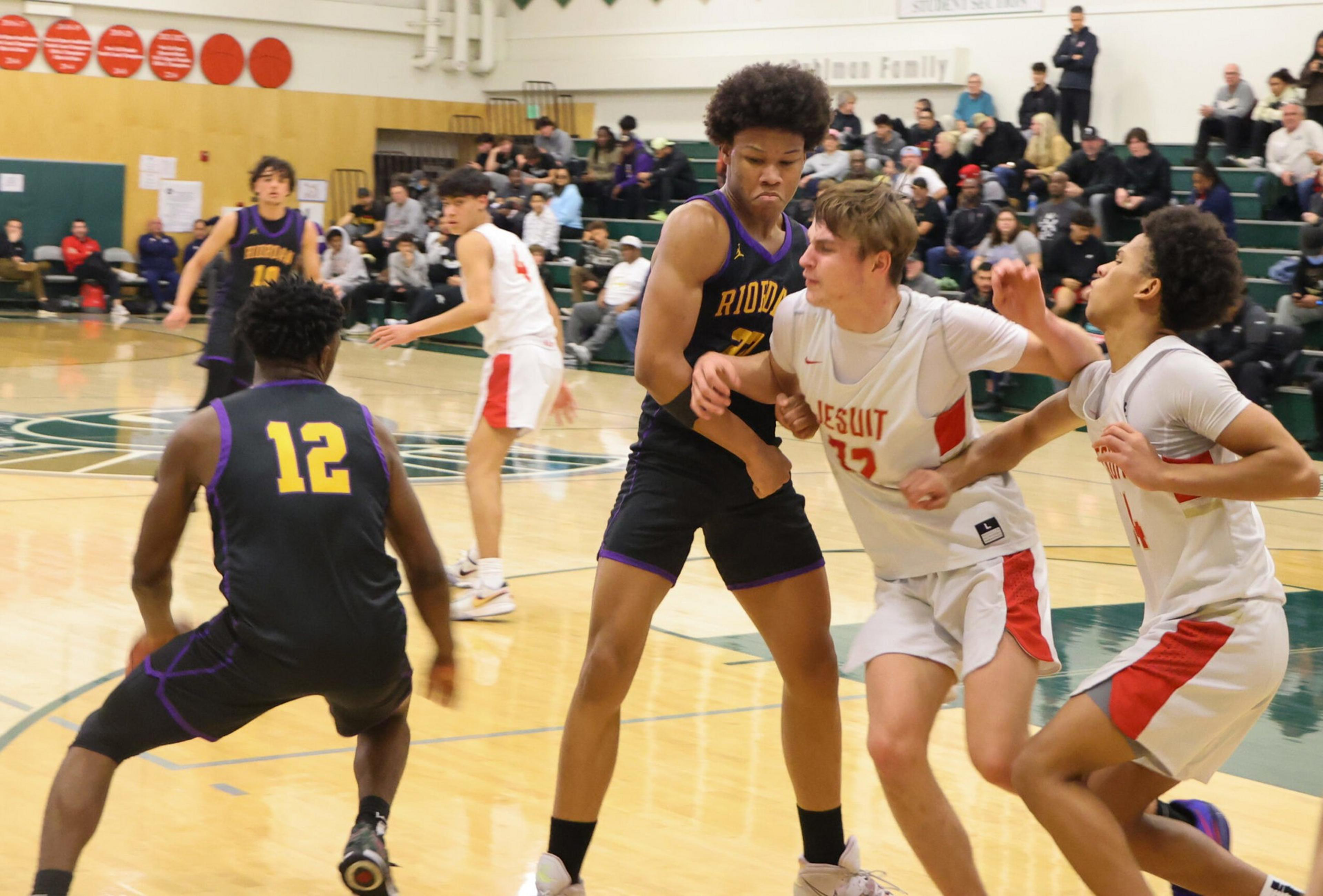 Two basketball players in white jerseys closely guard a player in a black and purple jersey, while another black and purple jersey player crouches nearby.
