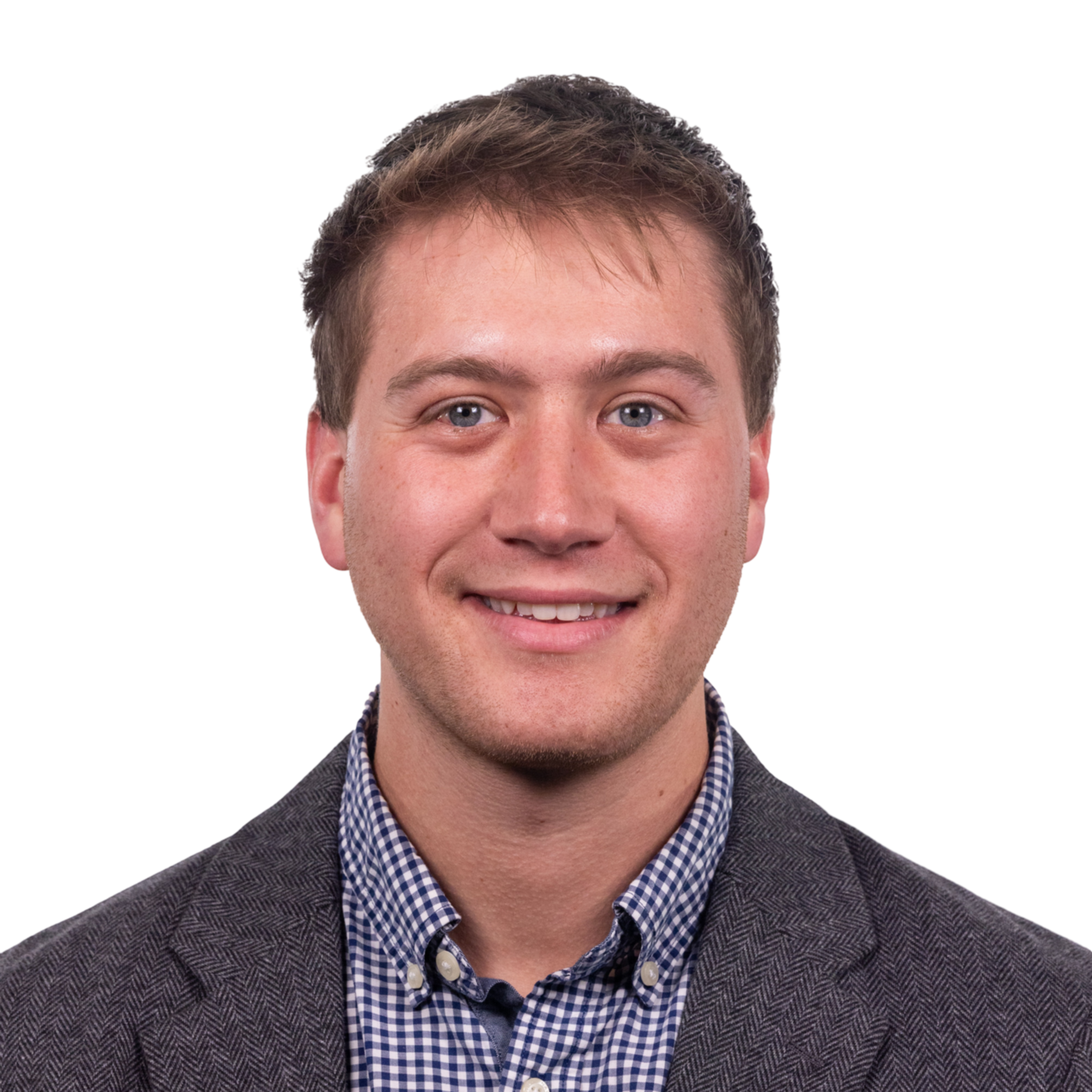 A young man with light skin and short brown hair wears a gray blazer over a blue and white checkered shirt, smiling directly at the camera.