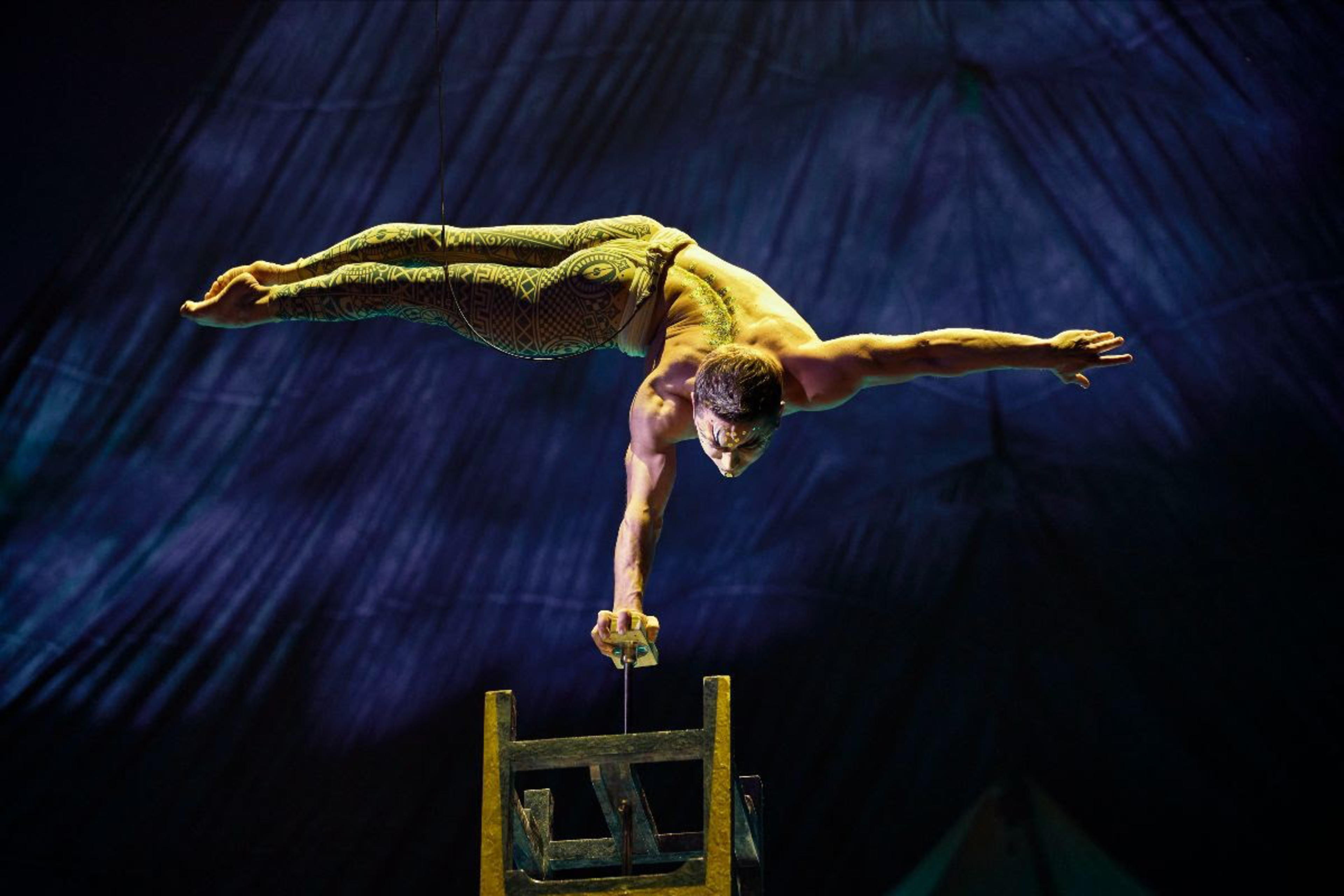 A shirtless acrobat balances horizontally on one hand atop a pole attached to a wooden chair, against a dark, stage-lit background.