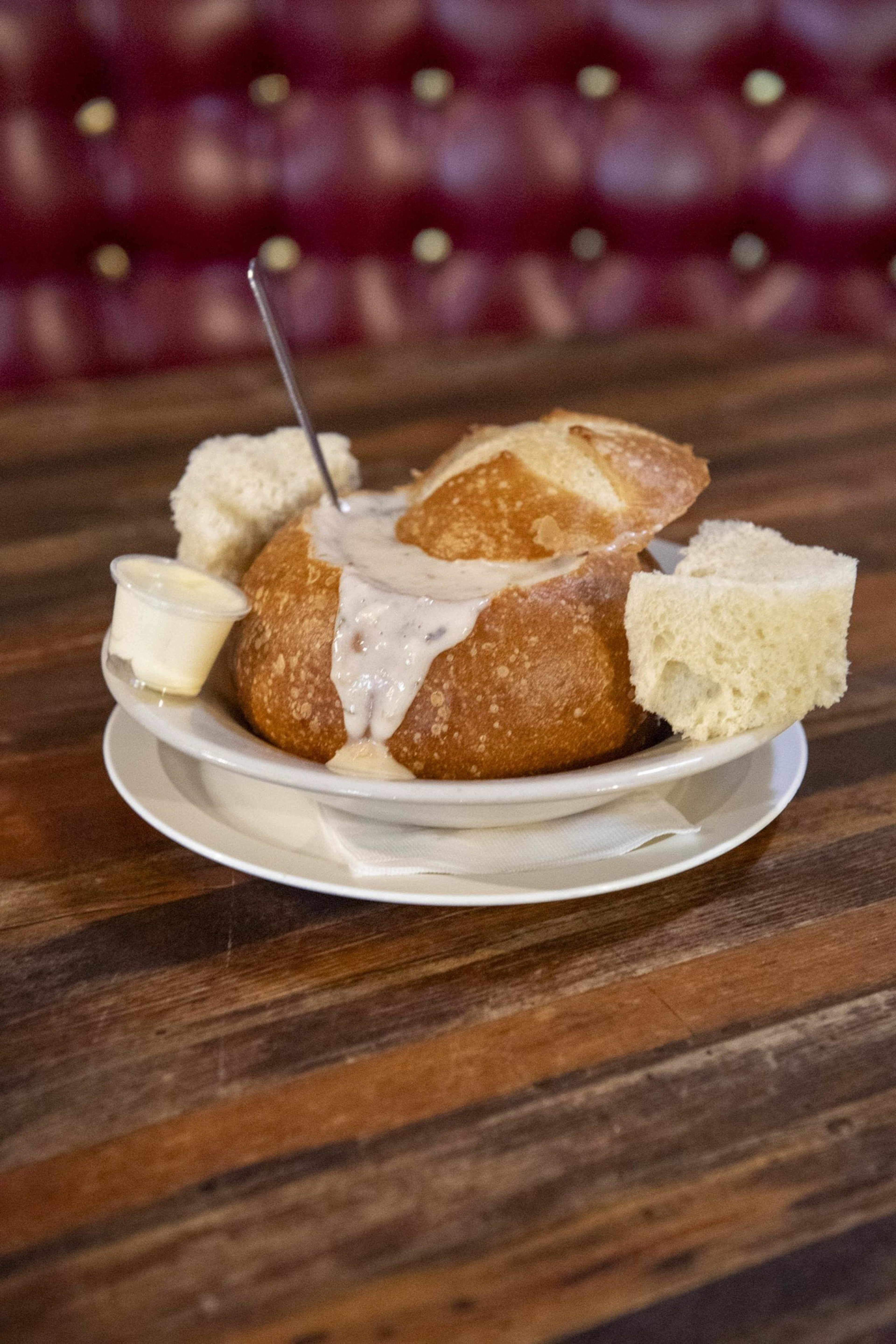 A bread bowl filled with creamy soup sits on a plate with bread pieces and a small container of butter. A spoon is placed in the soup.