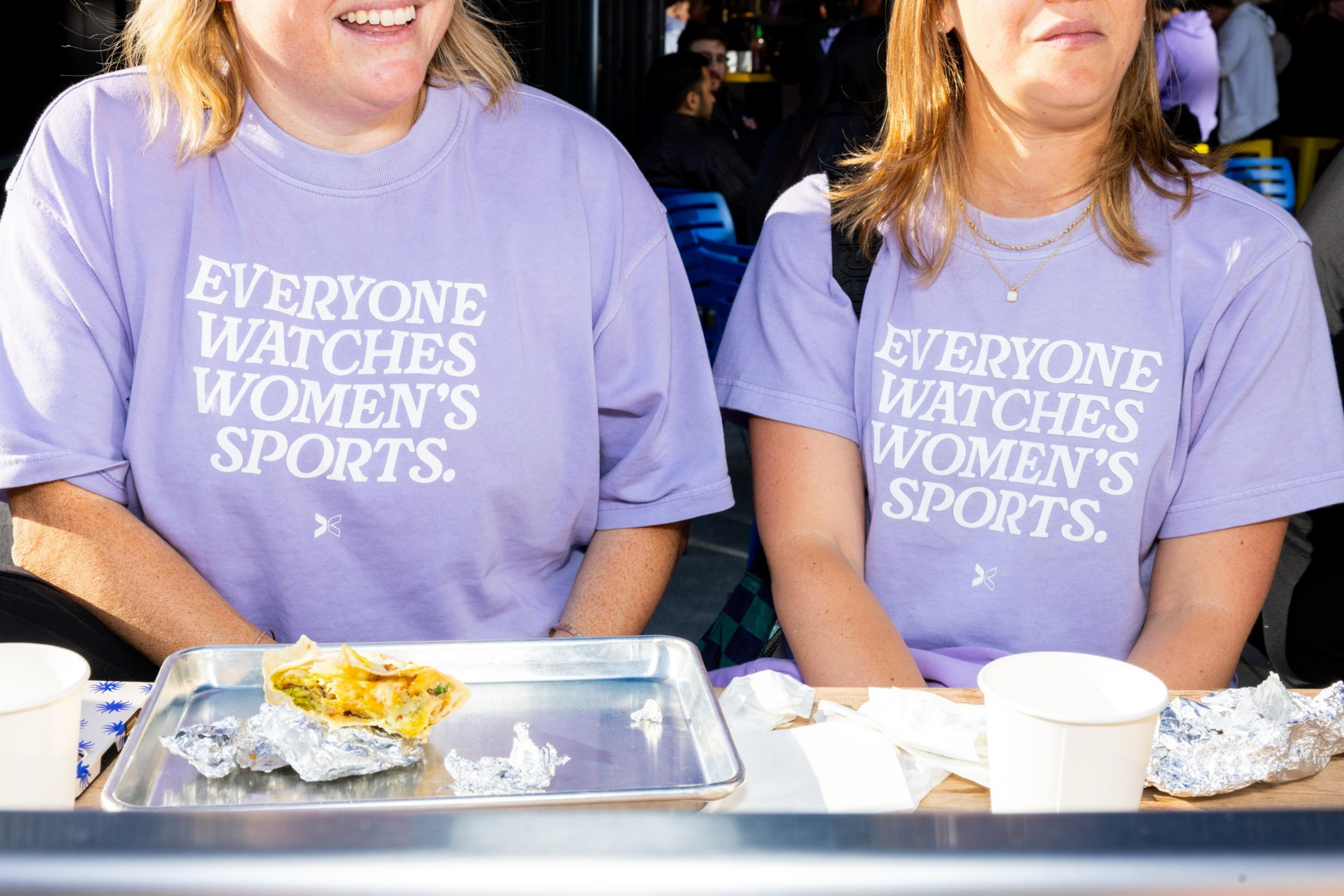 Two women wearing matching lavender shirts that say "EVERYONE WATCHES WOMEN'S SPORTS." sit at a table with trays, foil, and cups.
