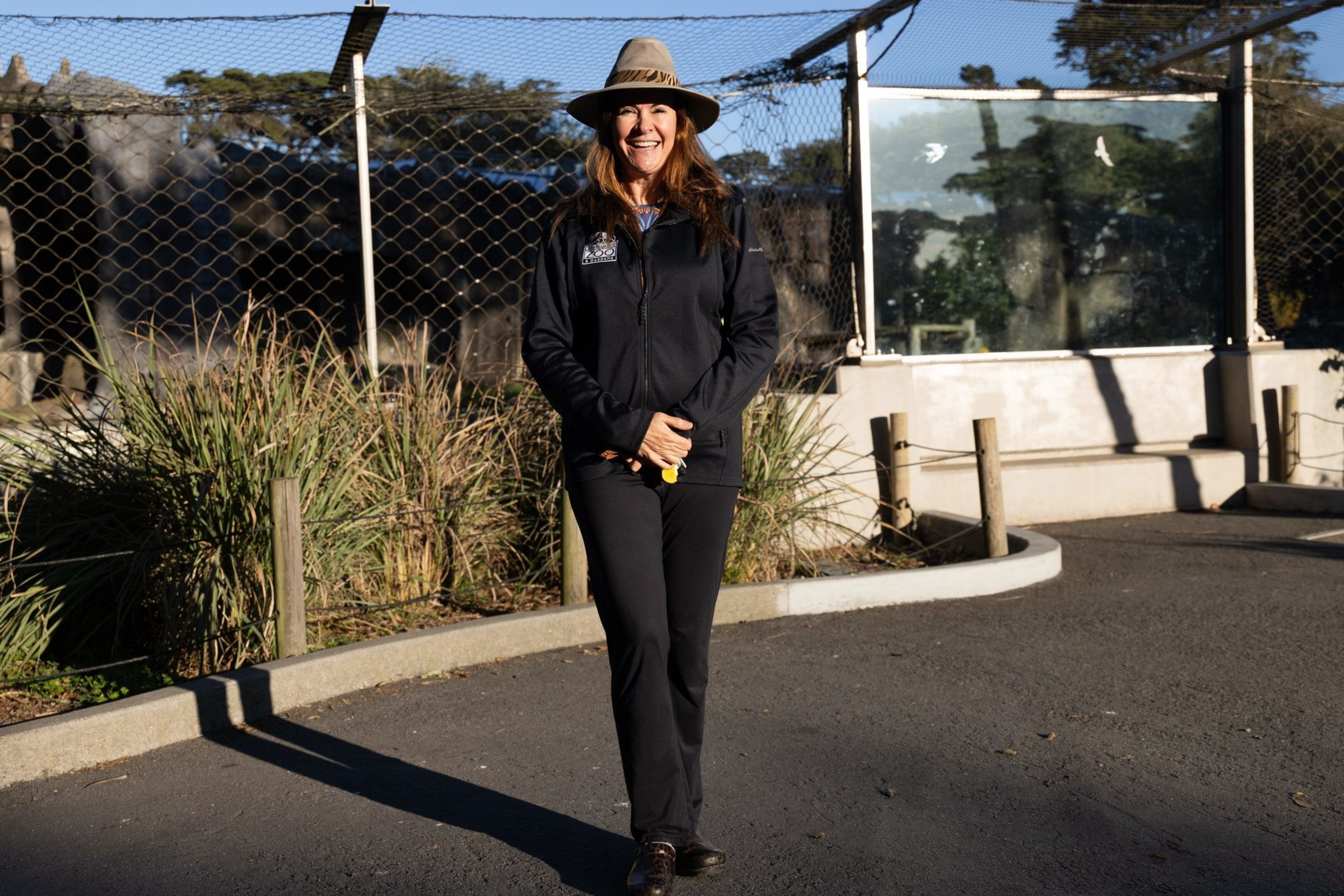 A smiling person stands on a paved path, wearing a wide-brimmed hat and dark clothing. They are in front of a fenced area with plants and trees.
