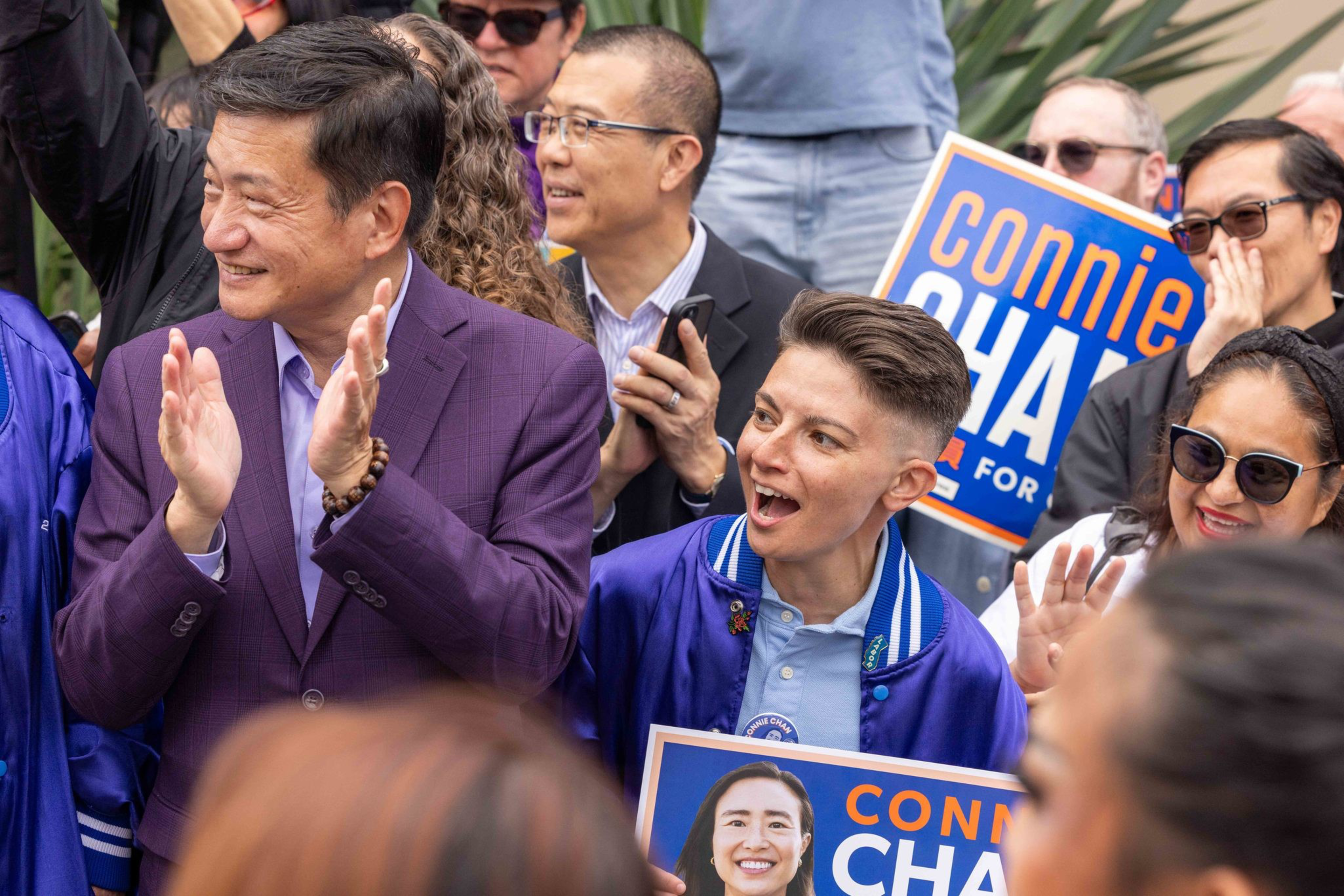 A group of diverse people cheer and hold campaign signs for Connie Chan at an outdoor event.