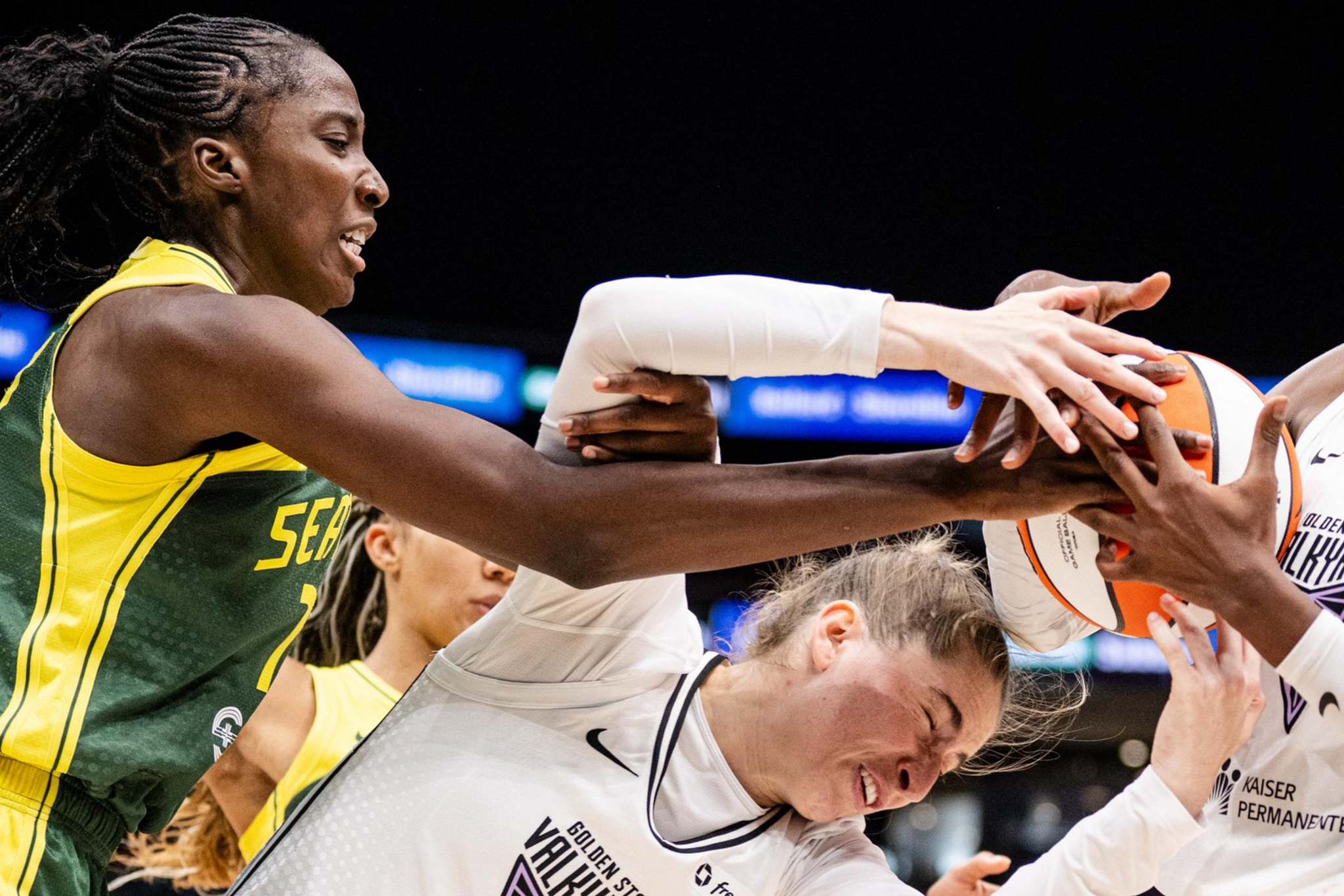 Three basketball players are vying intensely for control of the ball. Two players in white jerseys and one in a green jersey are in a tense struggle.