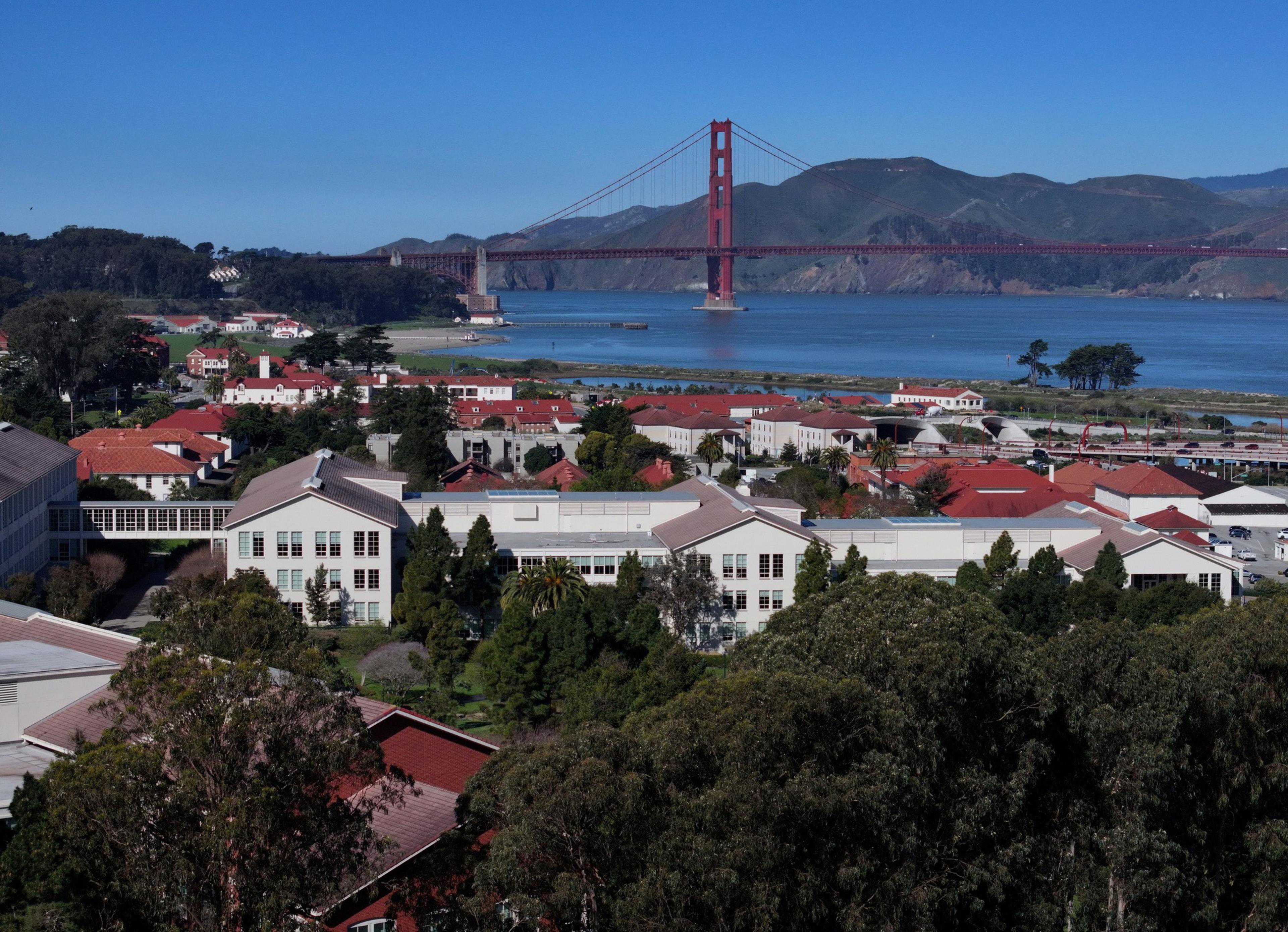 A coastal cityscape with red-roofed buildings, green trees, and the Golden Gate Bridge spanning across blue water under a clear sky.