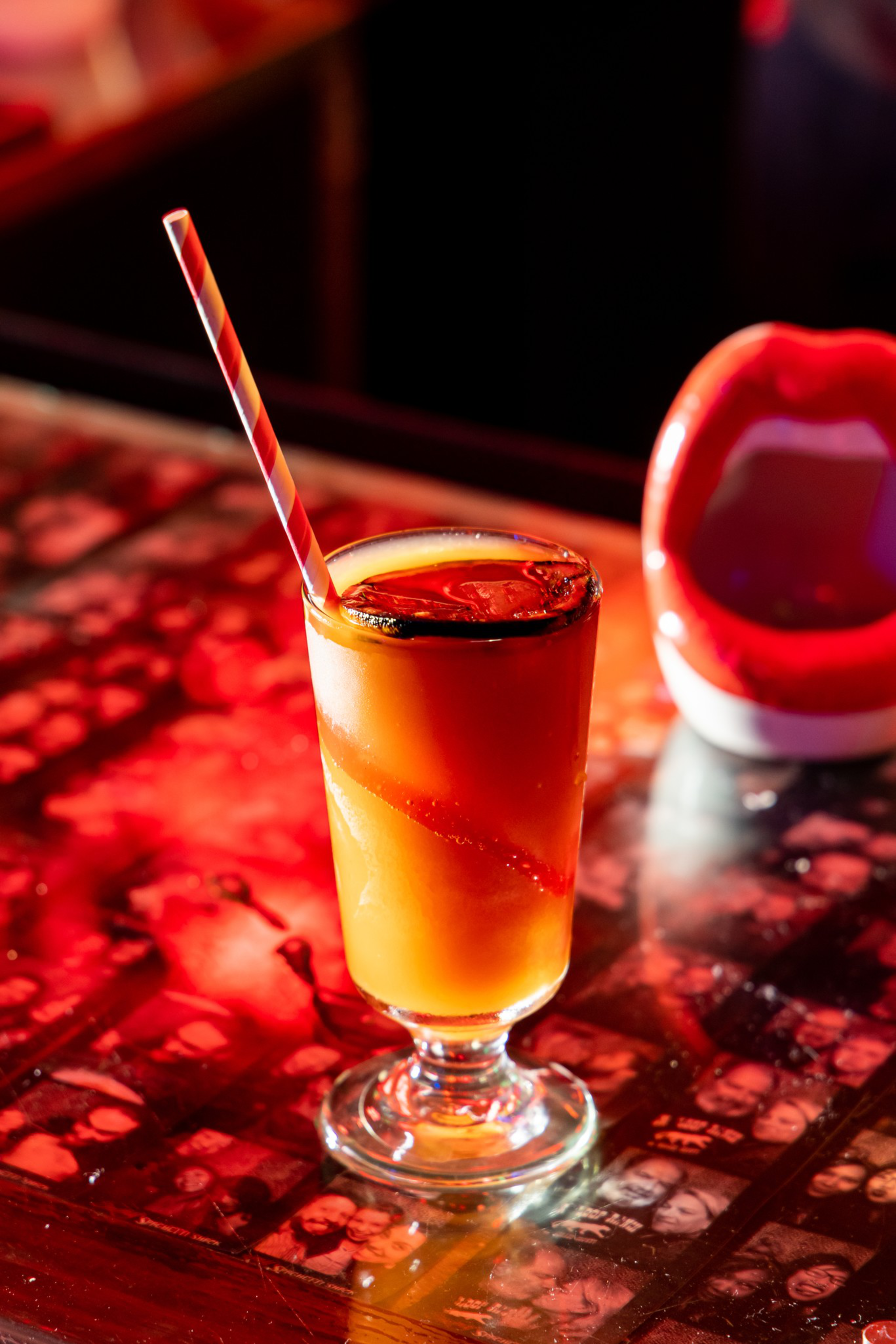 A tall glass of orange cocktail with ice and a red-and-white striped straw sits on a red-lit table covered with black-and-white photos.