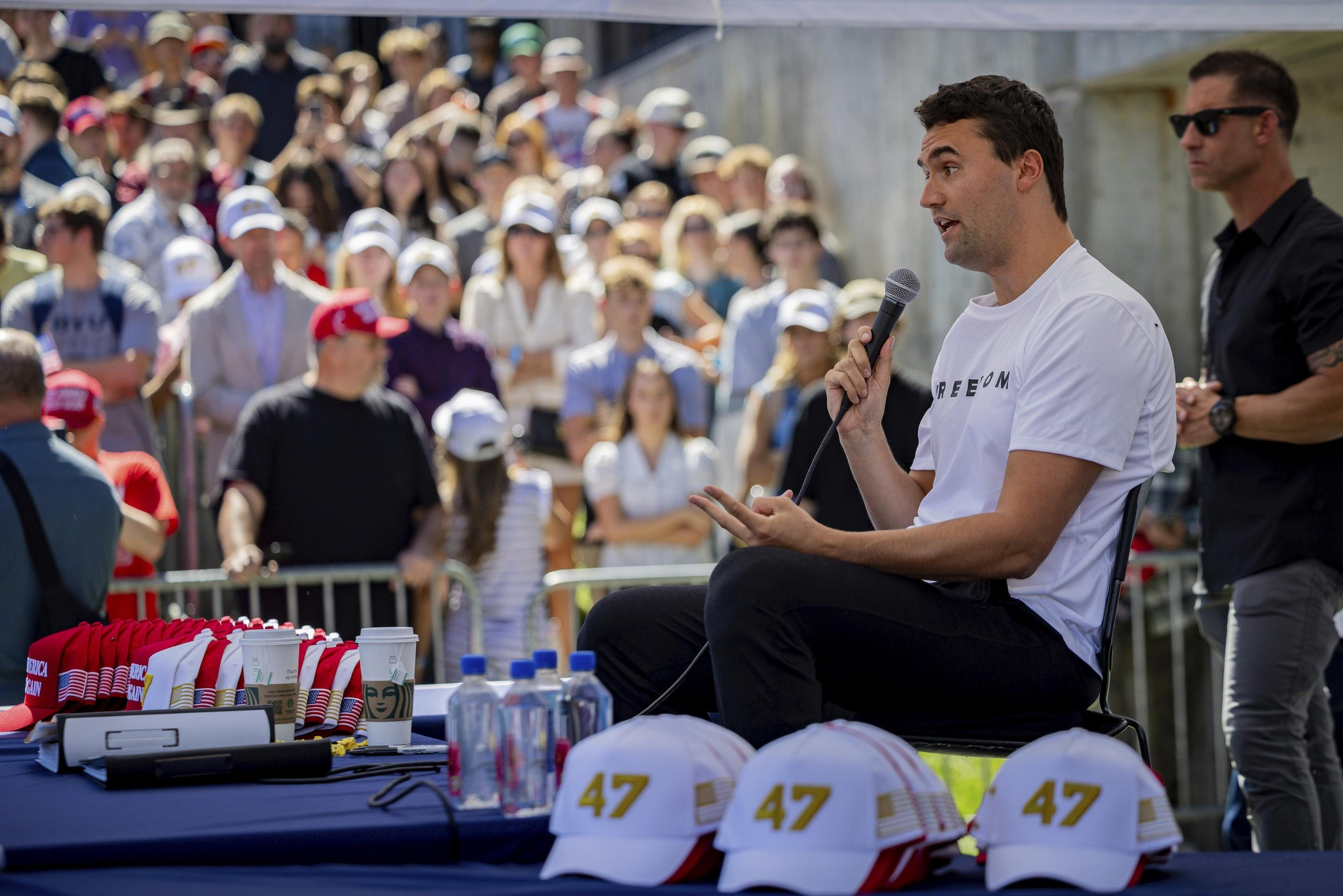 A man in a white shirt holding a microphone speaks to a crowd while seated, nearby hats displaying "47" and water bottles sit on a table.