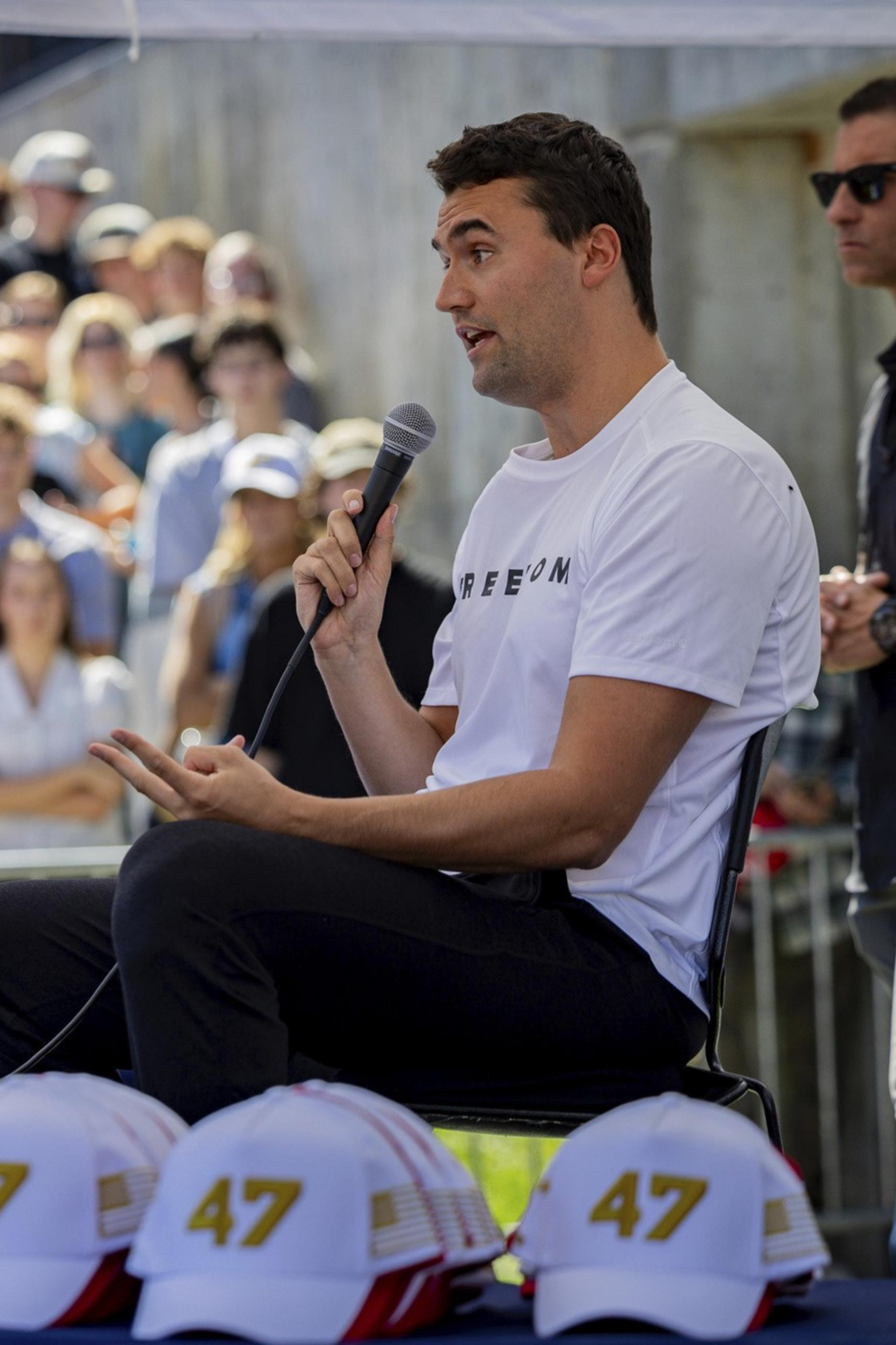 A man in a white shirt holding a microphone speaks to a crowd while seated, nearby hats displaying "47" and water bottles sit on a table.