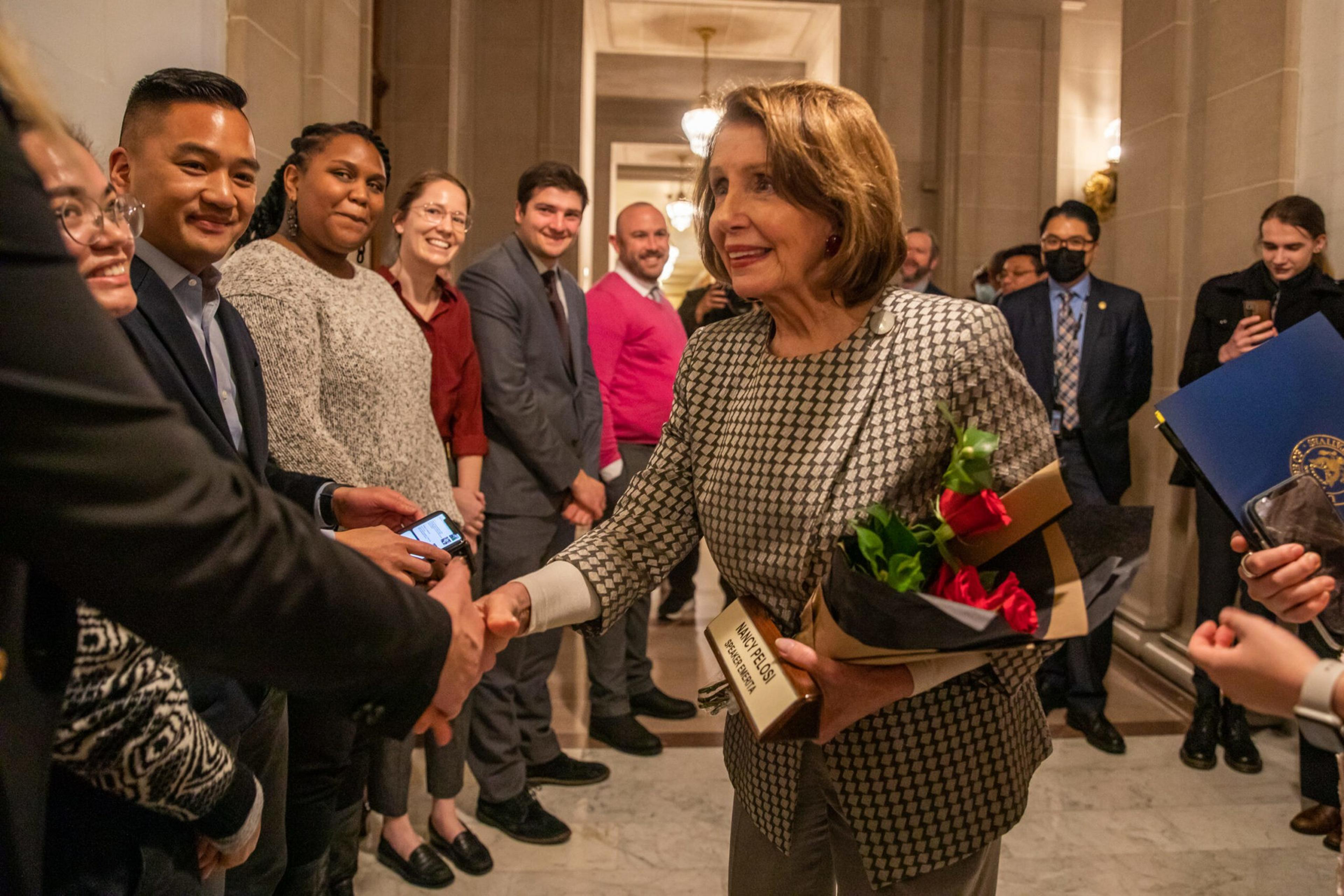 A woman holding flowers and a plaque shakes hands with smiling people lined up in a hallway, with some individuals wearing suits and one in a mask.