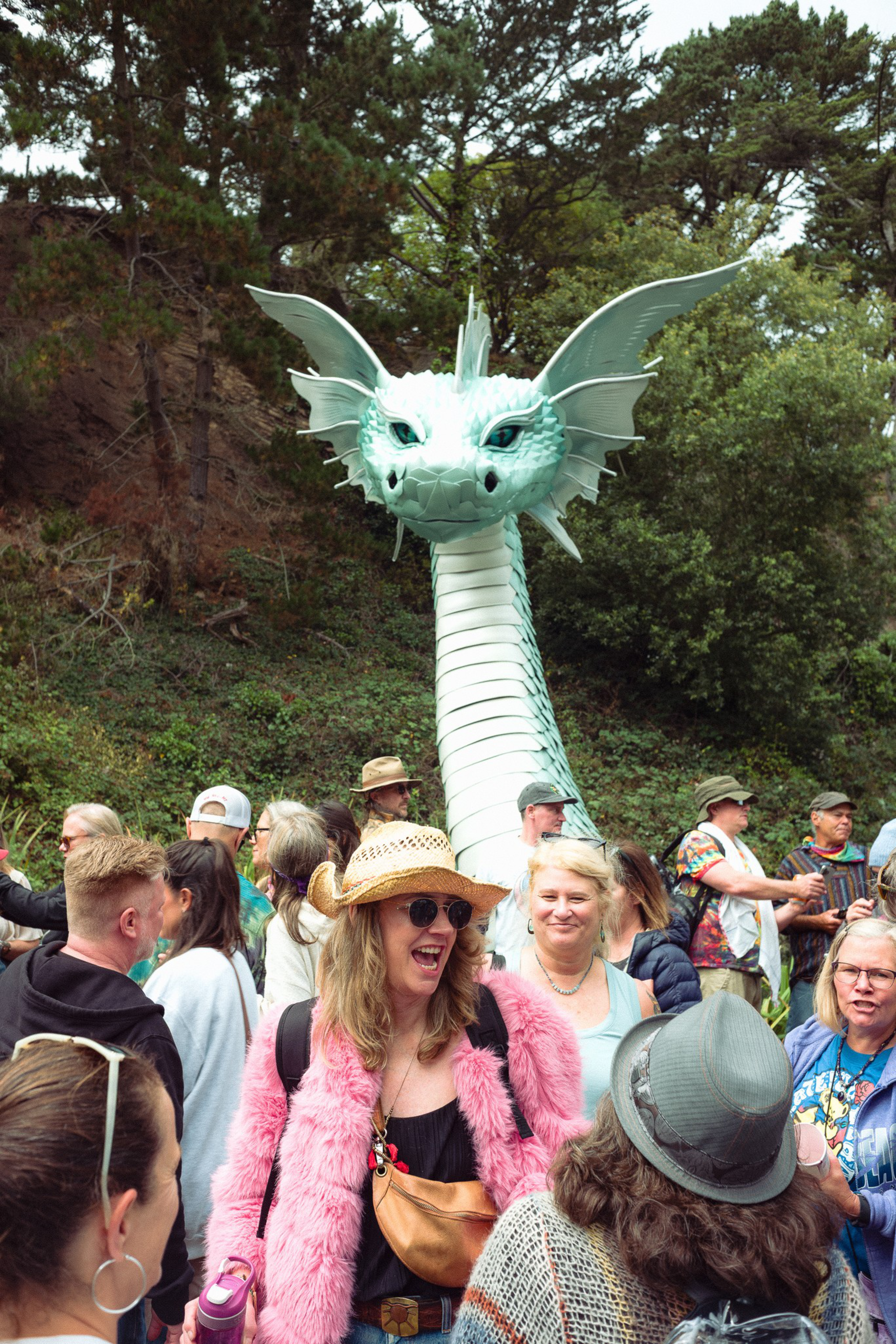 A large, pale green dragon sculpture with wings towers behind a crowd of smiling people outdoors near trees and bushes.
