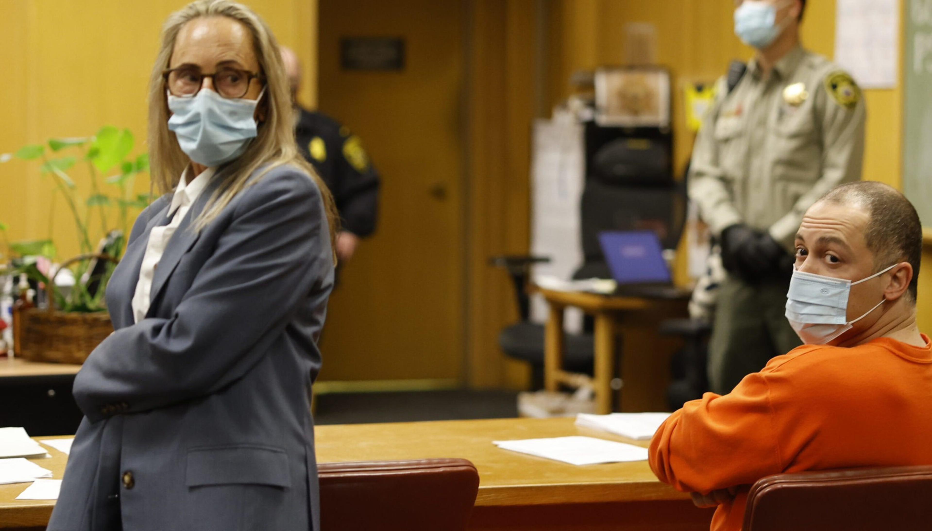 A man in an orange prison jumpsuit and a woman in a gray suit, both wearing face masks, sit in a courtroom with an officer standing in the background.